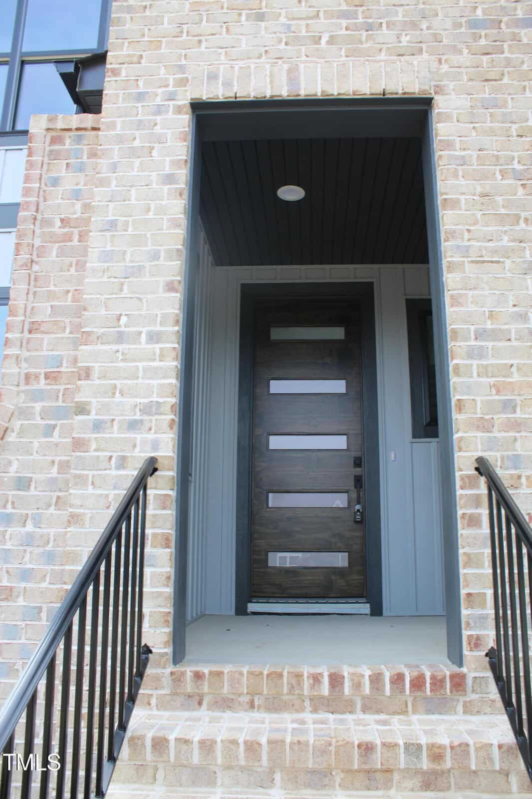 A front entrance with brick steps and black metal railings leads to a dark wooden door with four horizontal glass panels, set in a light-colored brick building.