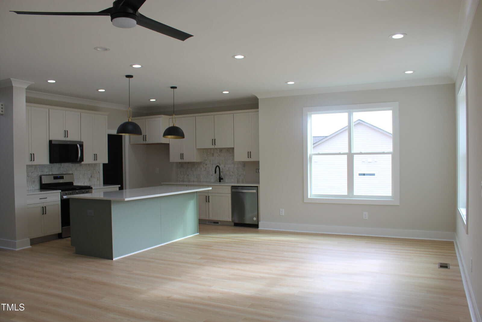 Modern open-concept kitchen with white cabinets, stainless steel appliances, and a large island with a light green base. Pendant lights hang above the island. Light wood flooring and large windows provide natural light.