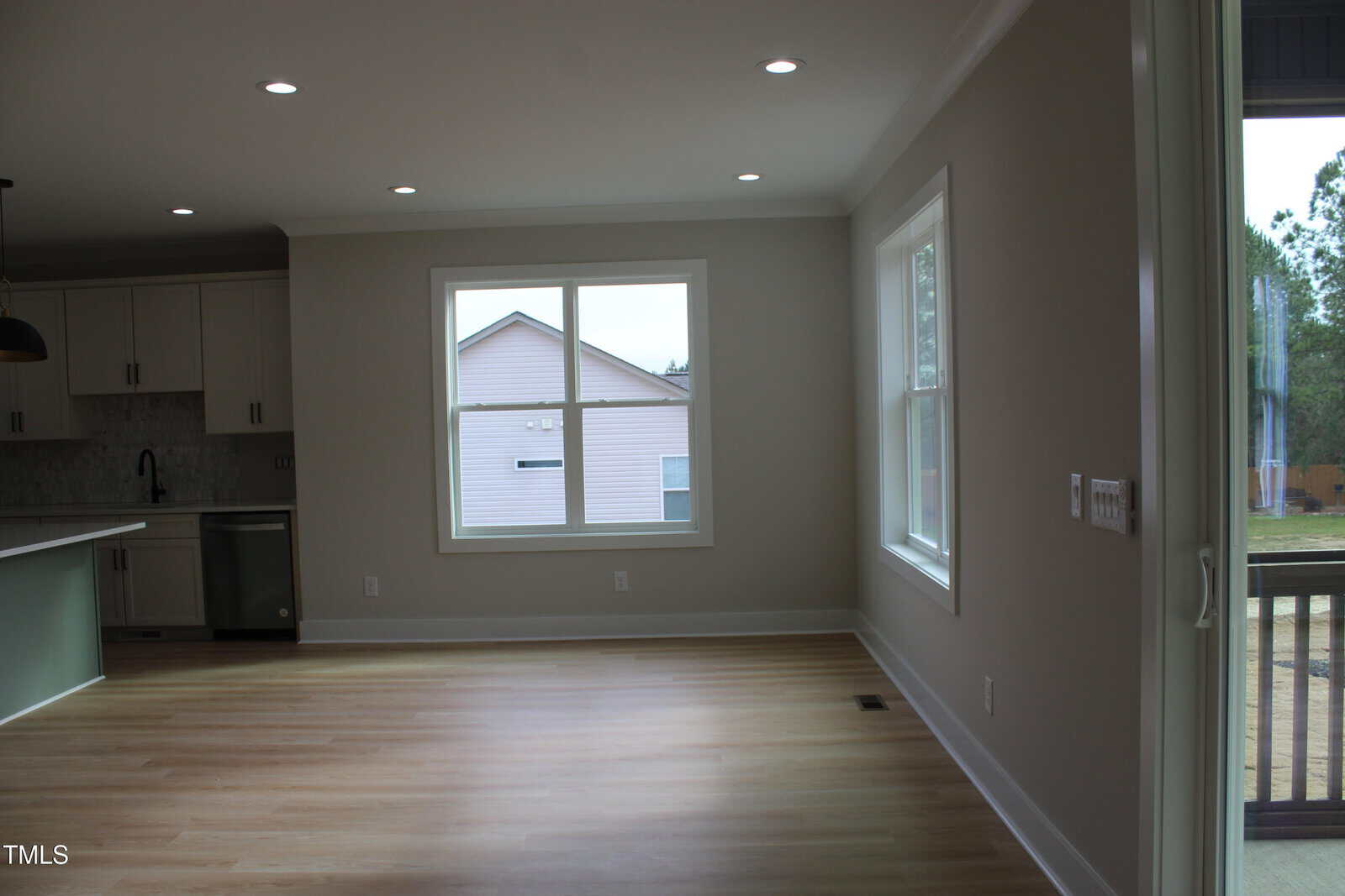A bright, empty room with light wood floors, beige walls, large windows, and recessed ceiling lights. Kitchen area with white cabinets and stainless steel appliances is visible on the left.