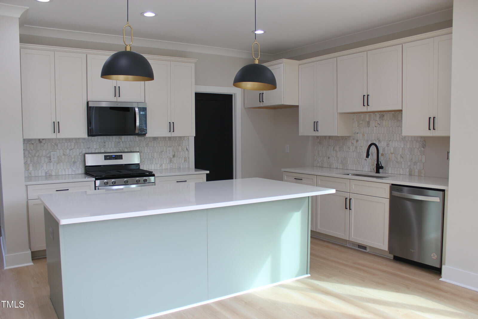 Modern kitchen with white cabinets, stainless steel appliances, a central island with a light green base, two black pendant lights, tile backsplash, light wood floors, and neutral walls.