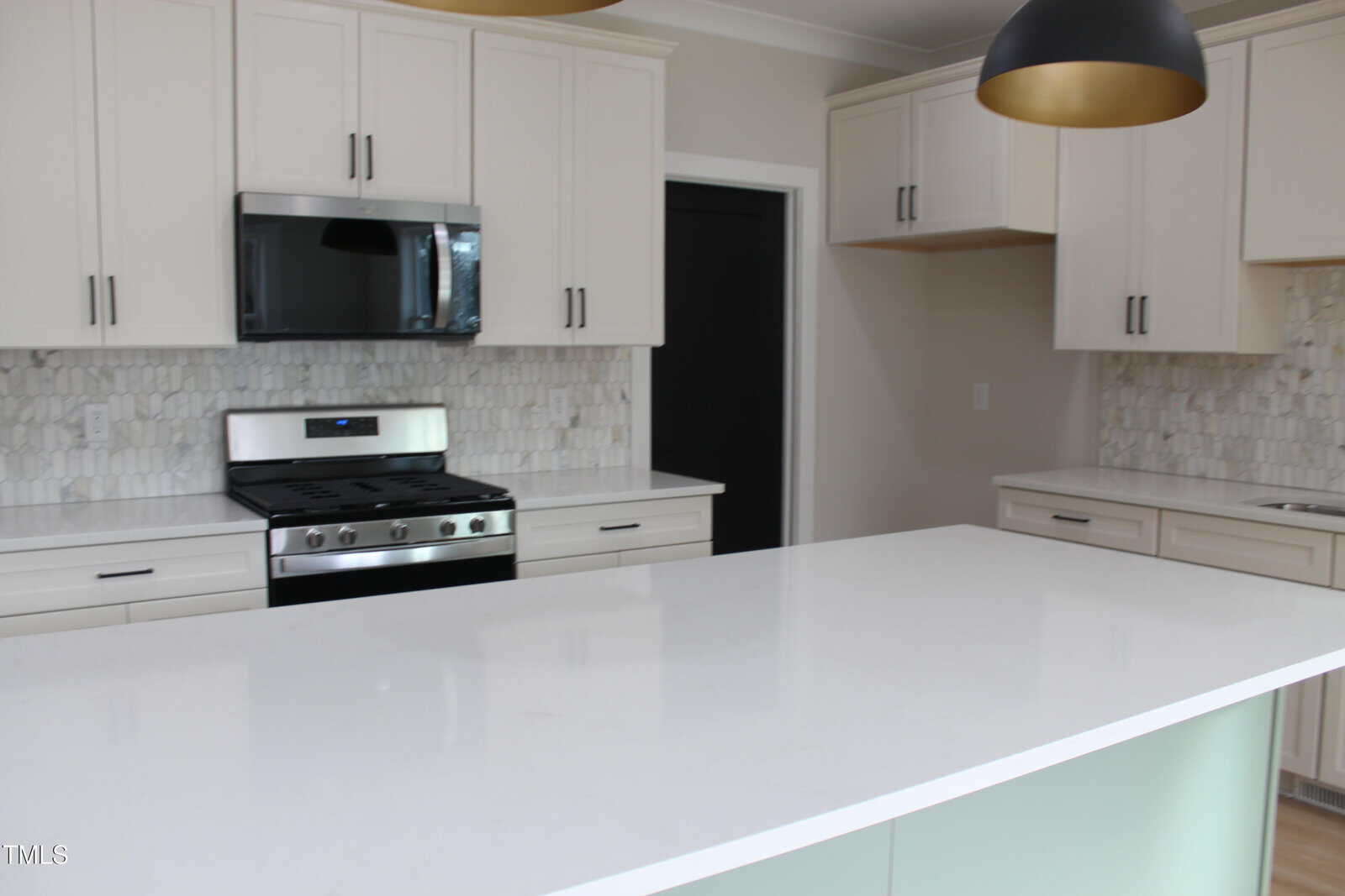 A modern kitchen with white cabinets, a white island countertop, a stainless steel gas stove and microwave, light-colored tile backsplash, and two black pendant lights.