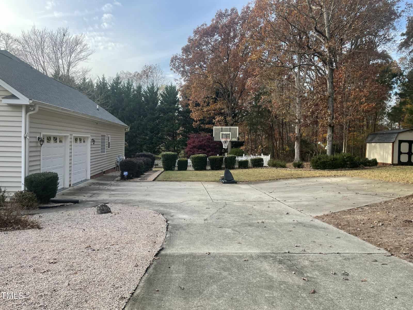 Concrete driveway with a portable basketball hoop, bordered by a garage on the left and trees with autumn foliage in the background; a small shed is visible on the right side.