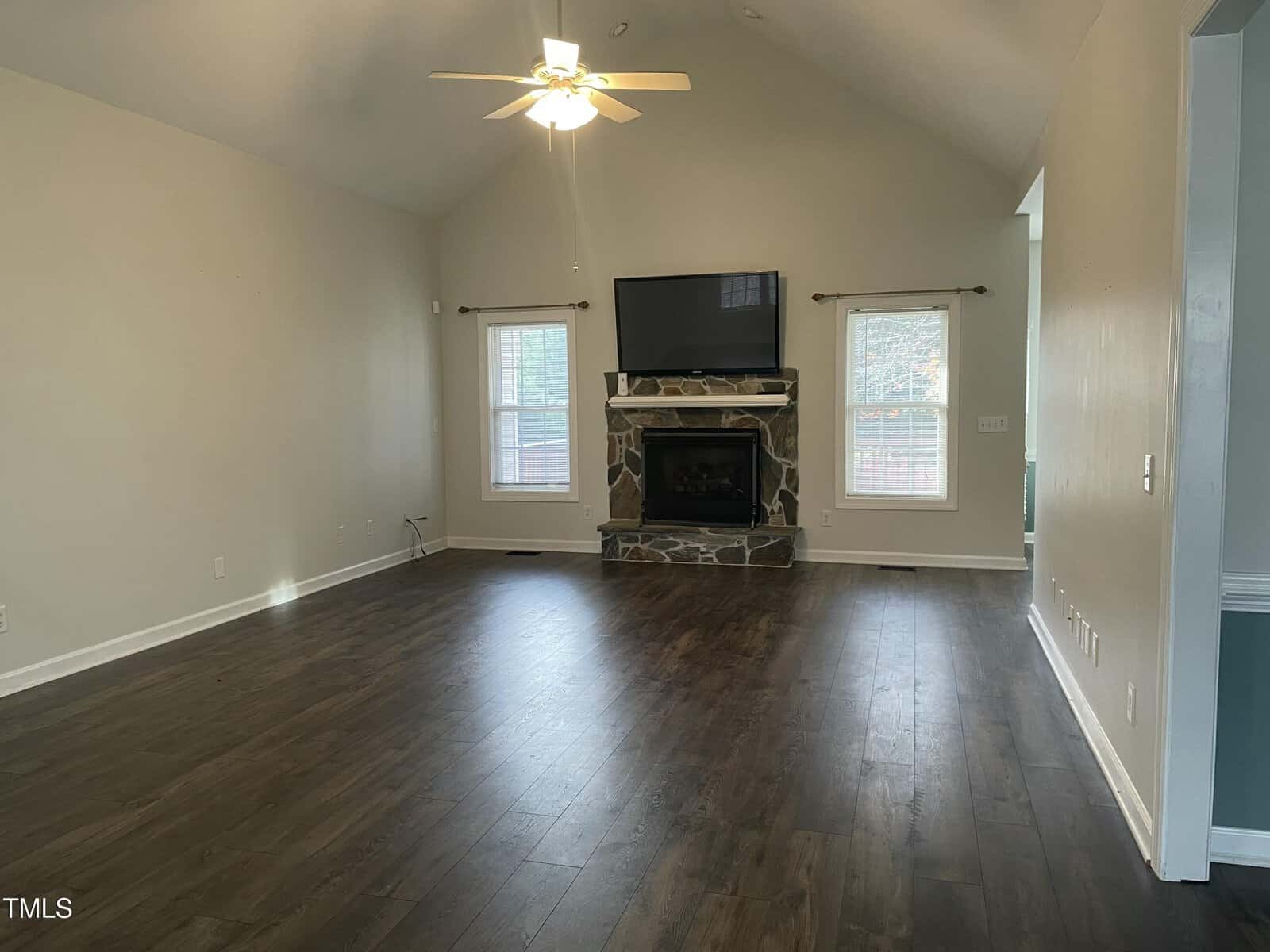 A spacious living room with dark wood floors, vaulted ceilings, light-colored walls, a ceiling fan, two windows, and a stone fireplace with a TV mounted above it. The room is unfurnished.