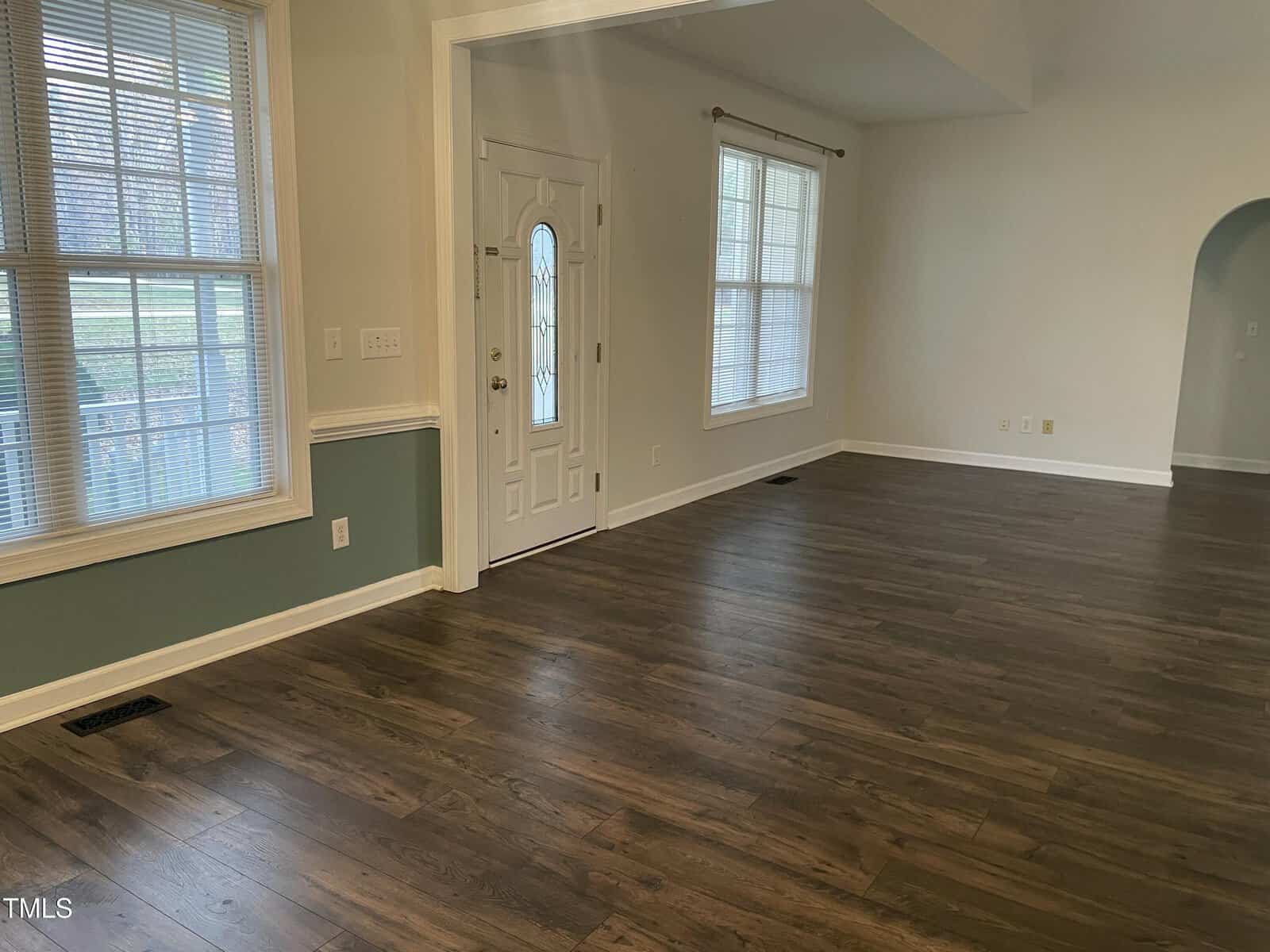Empty living room with wood flooring, large windows, white walls, and a white front door with a decorative glass panel. The space is well-lit with natural light from the windows.