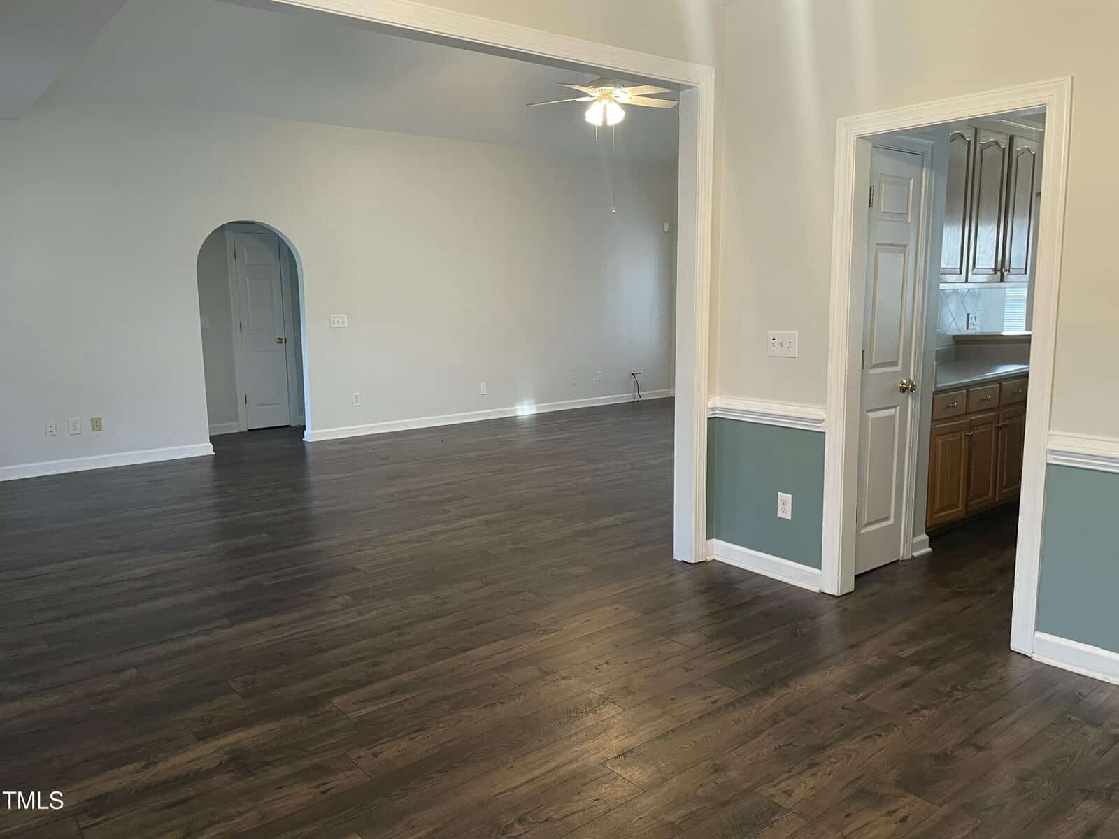 A spacious, empty room with dark wood floors, light-colored walls, a ceiling fan, and an open doorway leading to a kitchen with wooden cabinets. An arched doorway is visible in the background.