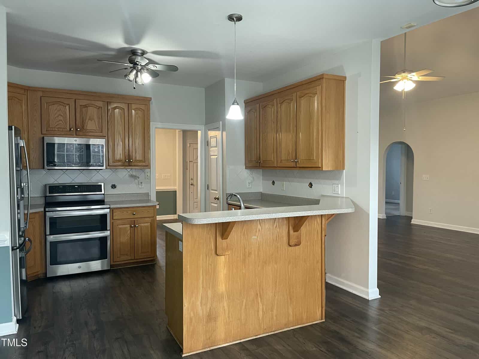 Modern kitchen with wooden cabinets, stainless steel appliances, a small island with a breakfast bar, and pendant lighting. Dark wood flooring throughout the kitchen and adjacent living area. Ceiling fans are visible in both spaces.