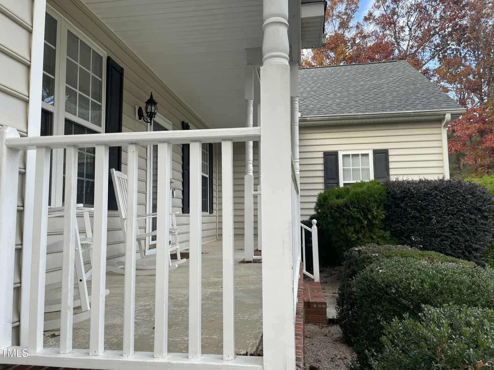A beige house with white trim features a covered front porch, two white rocking chairs, white railings, and a black lantern-style light fixture. Shrubs and brick steps lead up to the porch.