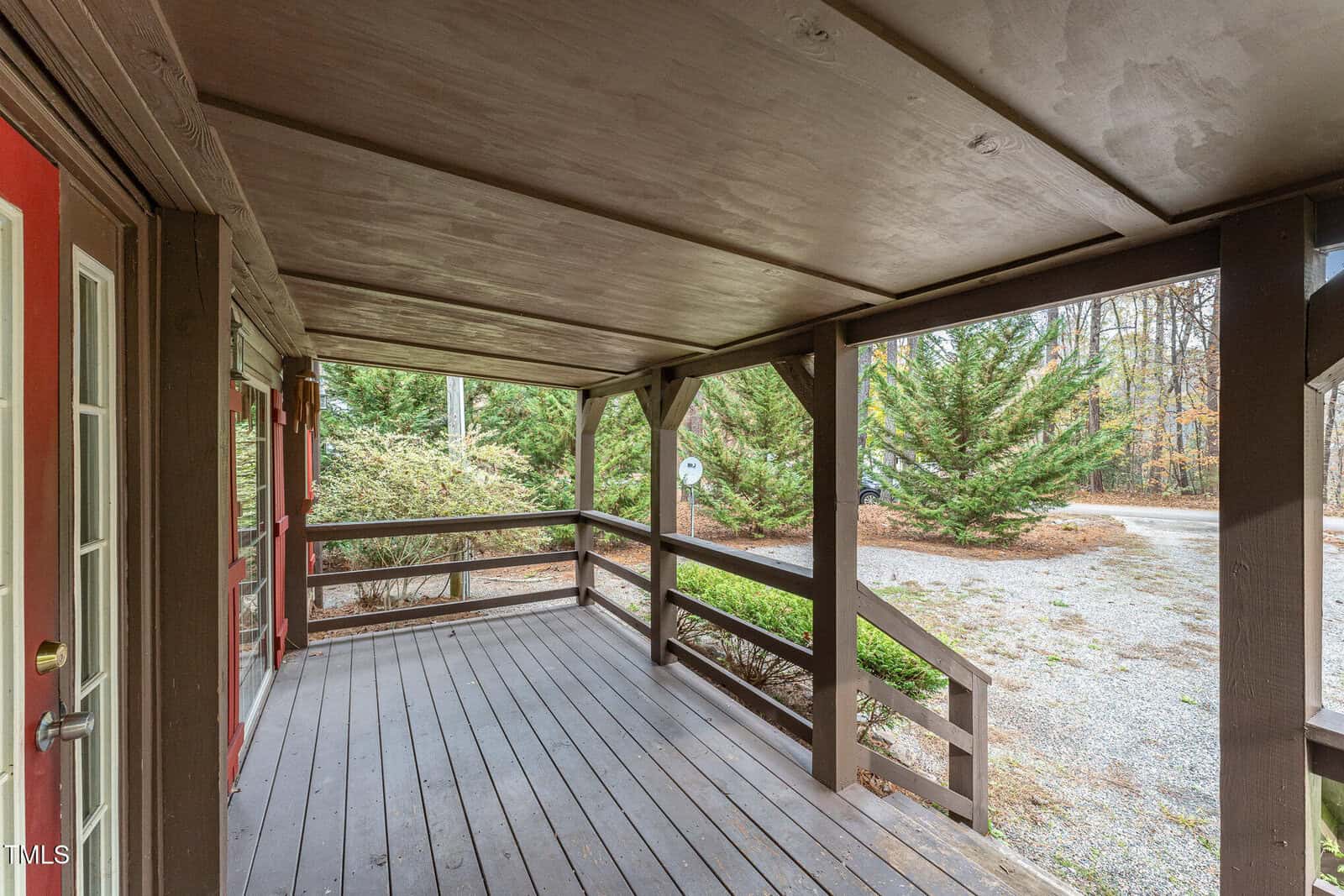 Covered wooden front porch with brown railings and beams, looking out toward a gravel driveway, green trees, and a wooded background. A red front door is visible on the left side.