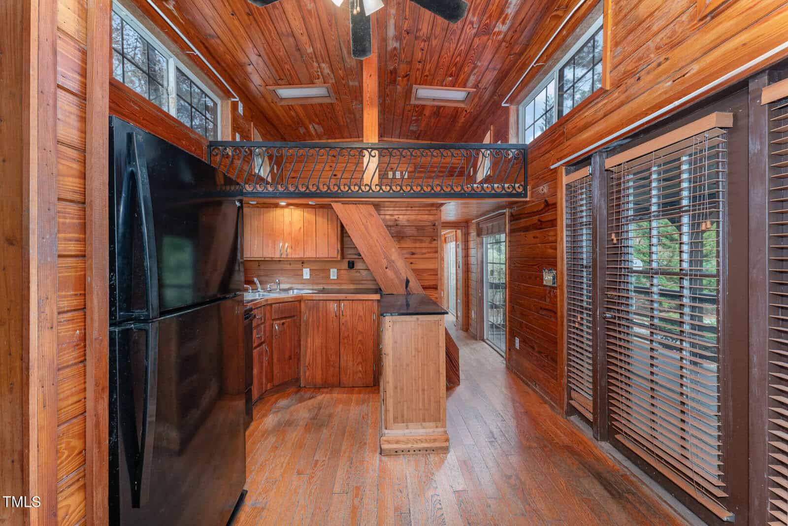 Kitchen with wood floors, walls, and ceiling, black refrigerator, wood cabinets, a loft-style railing above, and large windows with blinds letting in natural light. Stairs are visible in the center of the image.