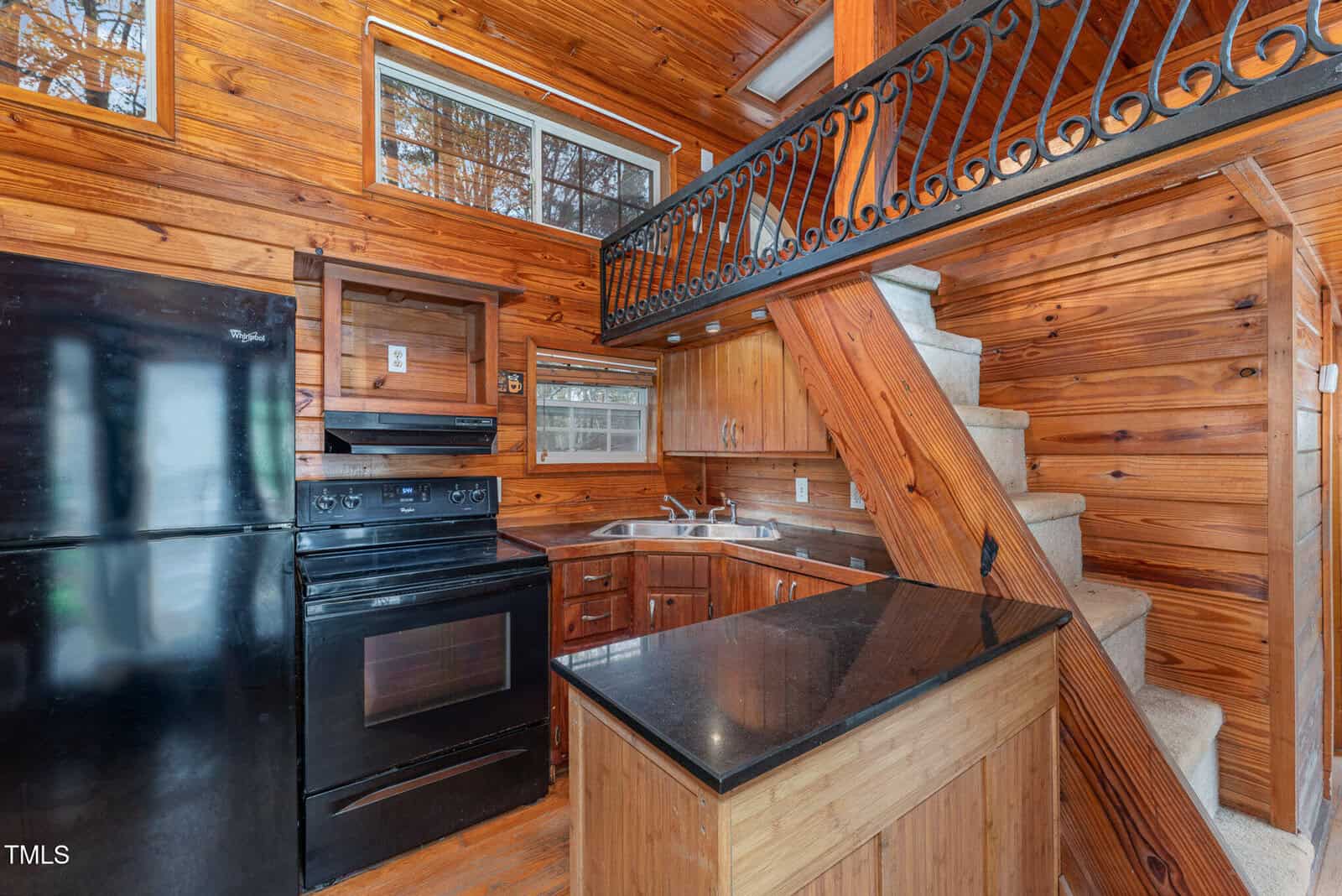 Compact wooden kitchen with black appliances, a loft above, and a staircase with a decorative black railing. Upper window and under-stairs cabinet add to the cozy, rustic atmosphere.