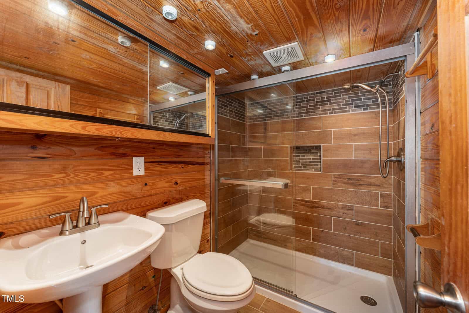 A bathroom with wood-paneled walls and ceiling, a white pedestal sink, a white toilet, and a walk-in glass shower with brown tile walls and a built-in shelf. Recessed lights are installed in the ceiling.