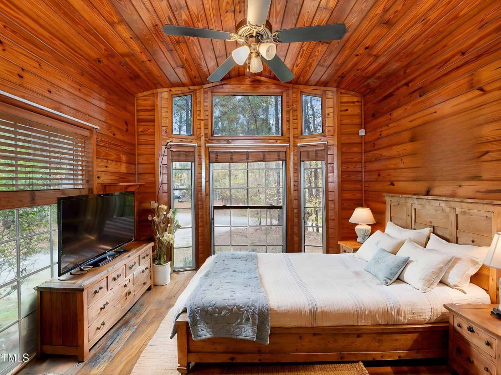 A bedroom with wood-paneled walls and ceiling, a large bed with white linens, a ceiling fan, a dresser with a TV, tall windows with a view of trees, and a small bedside lamp.