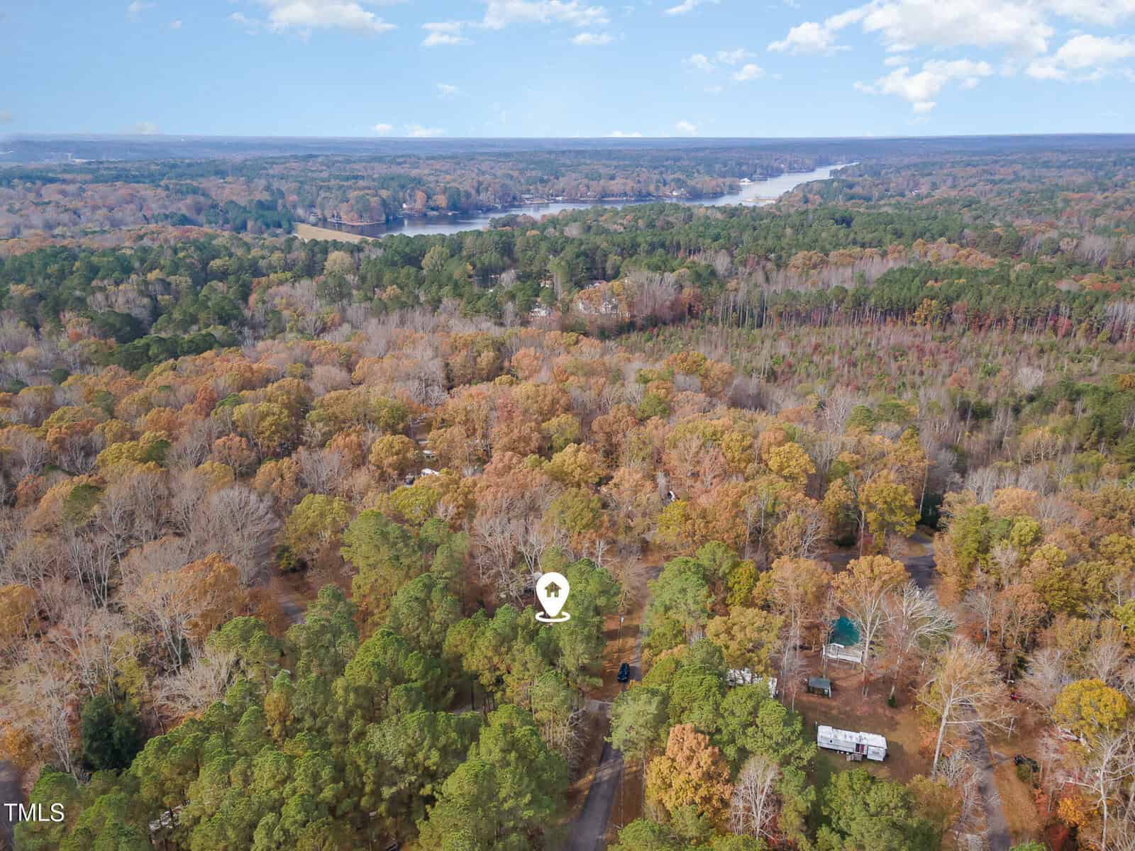 Aerial view of a wooded area with a mix of green and autumn-colored trees, scattered houses, a road, and a lake in the distance under a partly cloudy sky. A location marker highlights a spot among the trees.