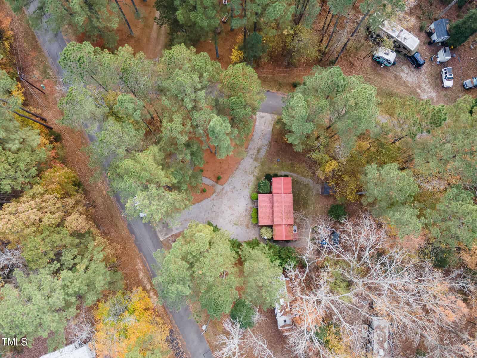 Aerial view of a red-roofed house surrounded by tall trees and a curved driveway, with nearby parked vehicles and scattered autumn leaves visible.