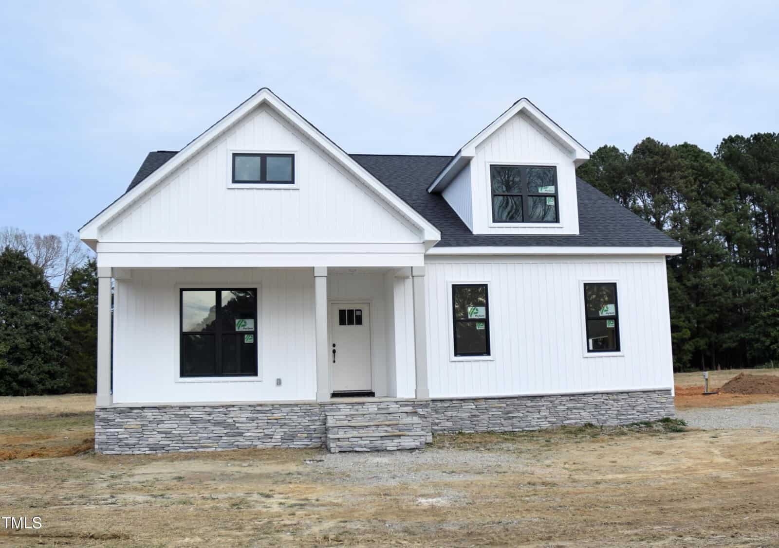 A newly built white house with a covered front porch, dark roof, stone foundation, and several windows. The yard is unfinished, with bare ground and some grass. Tall trees are visible in the background.