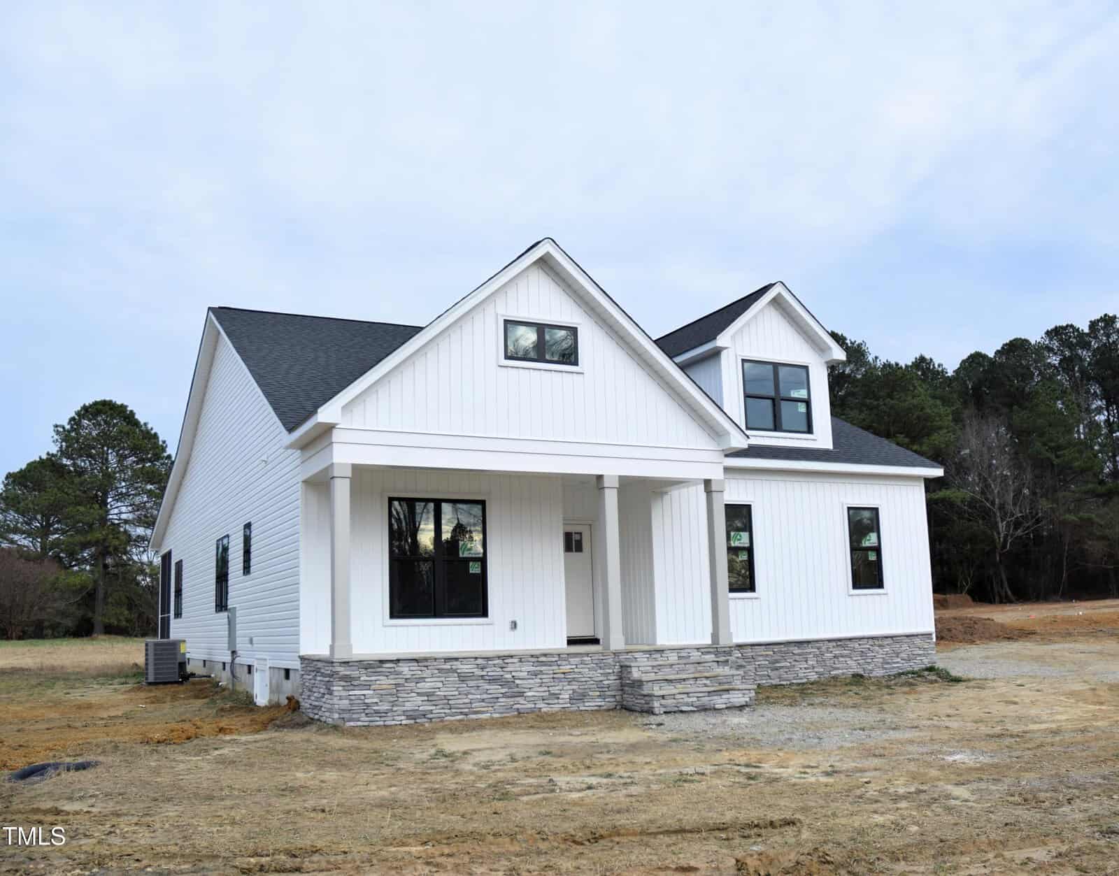 A newly constructed white house with a dark gray roof, featuring large windows and a stone foundation, sits on a bare dirt lot with some grass and trees in the background.