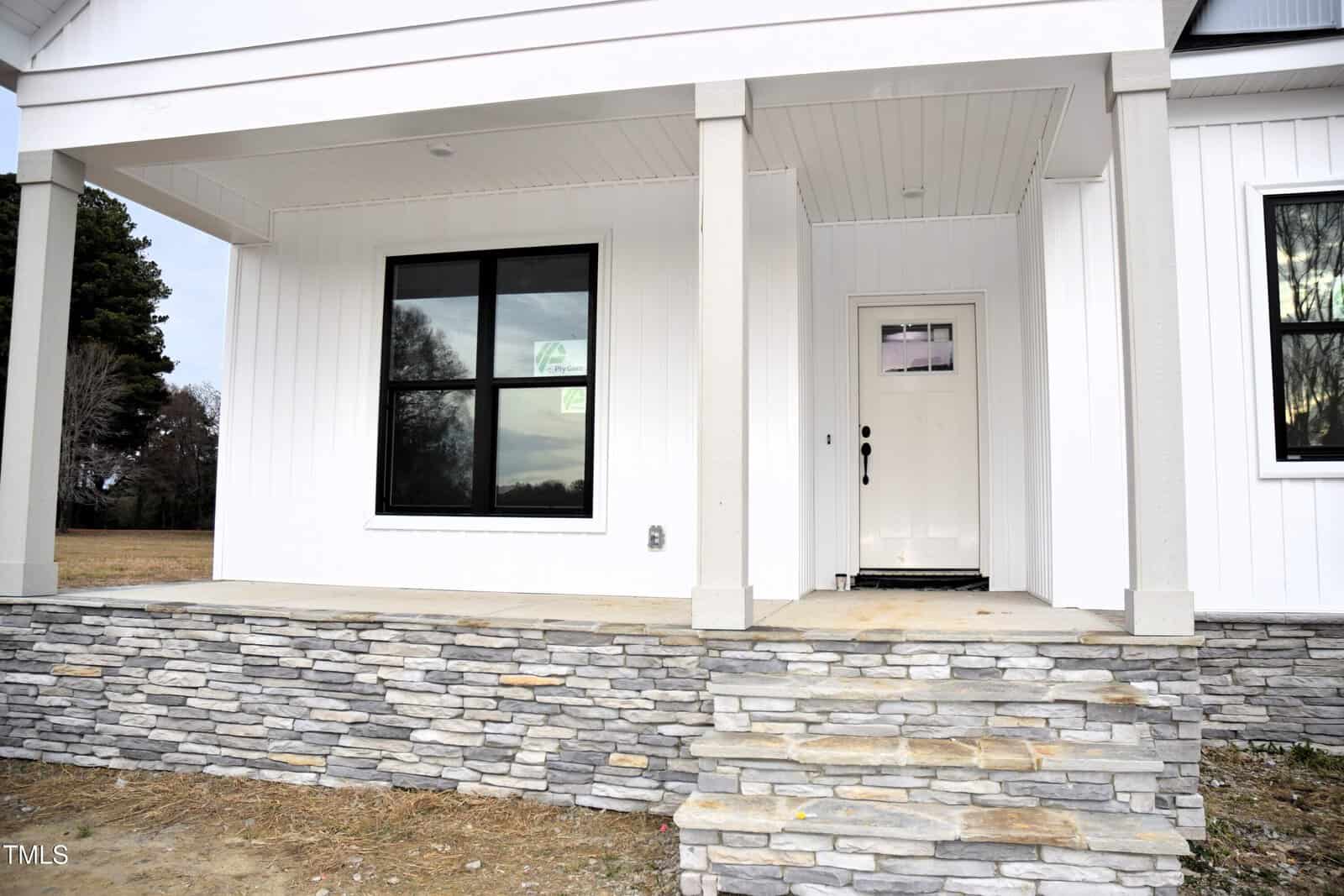 A modern house front with stone steps leading to a covered porch, white exterior walls, black-framed window, and a white door with glass panes. The yard is unfinished with bare ground and some grass.