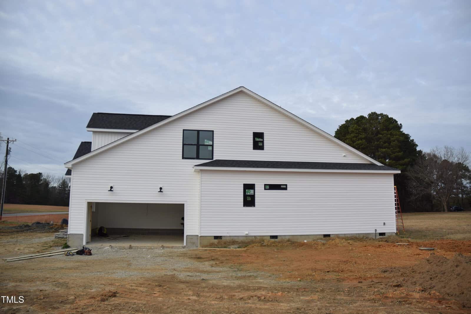 A newly constructed white house with black-trimmed windows and an open garage, surrounded by bare dirt and grass under a cloudy sky. Construction materials and a ladder are visible near the building.