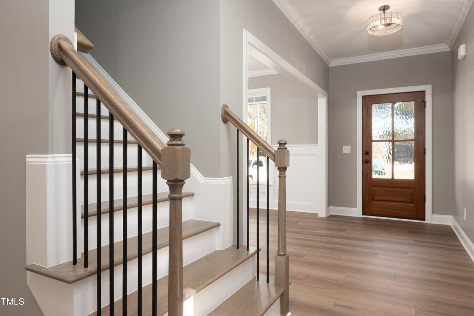 Modern home entryway with wooden stairs, black railings, light gray walls, crown molding, hardwood floors, a decorative light fixture, and a wooden front door with glass panels. Sunlight enters from a nearby window.