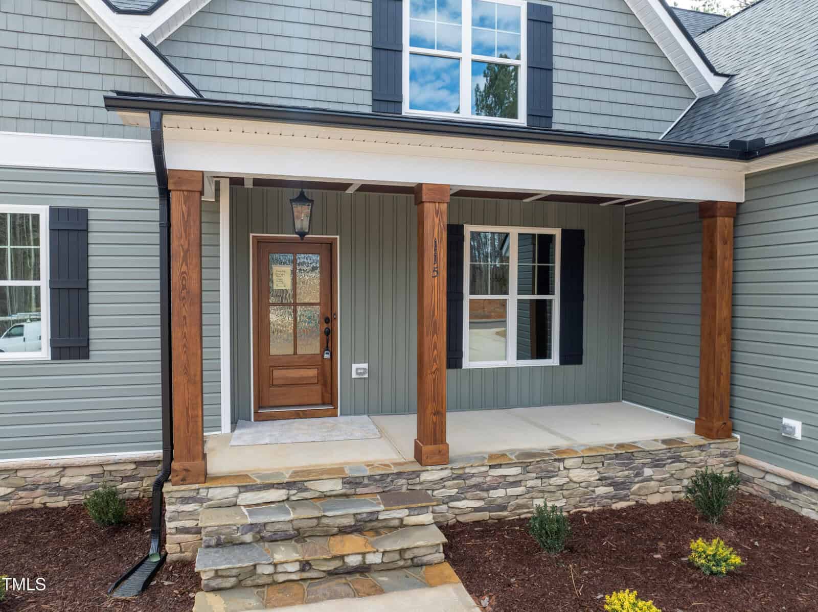 A front porch of a house with stone steps, wooden columns, a brown front door with glass panels, gray siding, black shutters, and a window above the entrance. Mulched planting beds are in front.