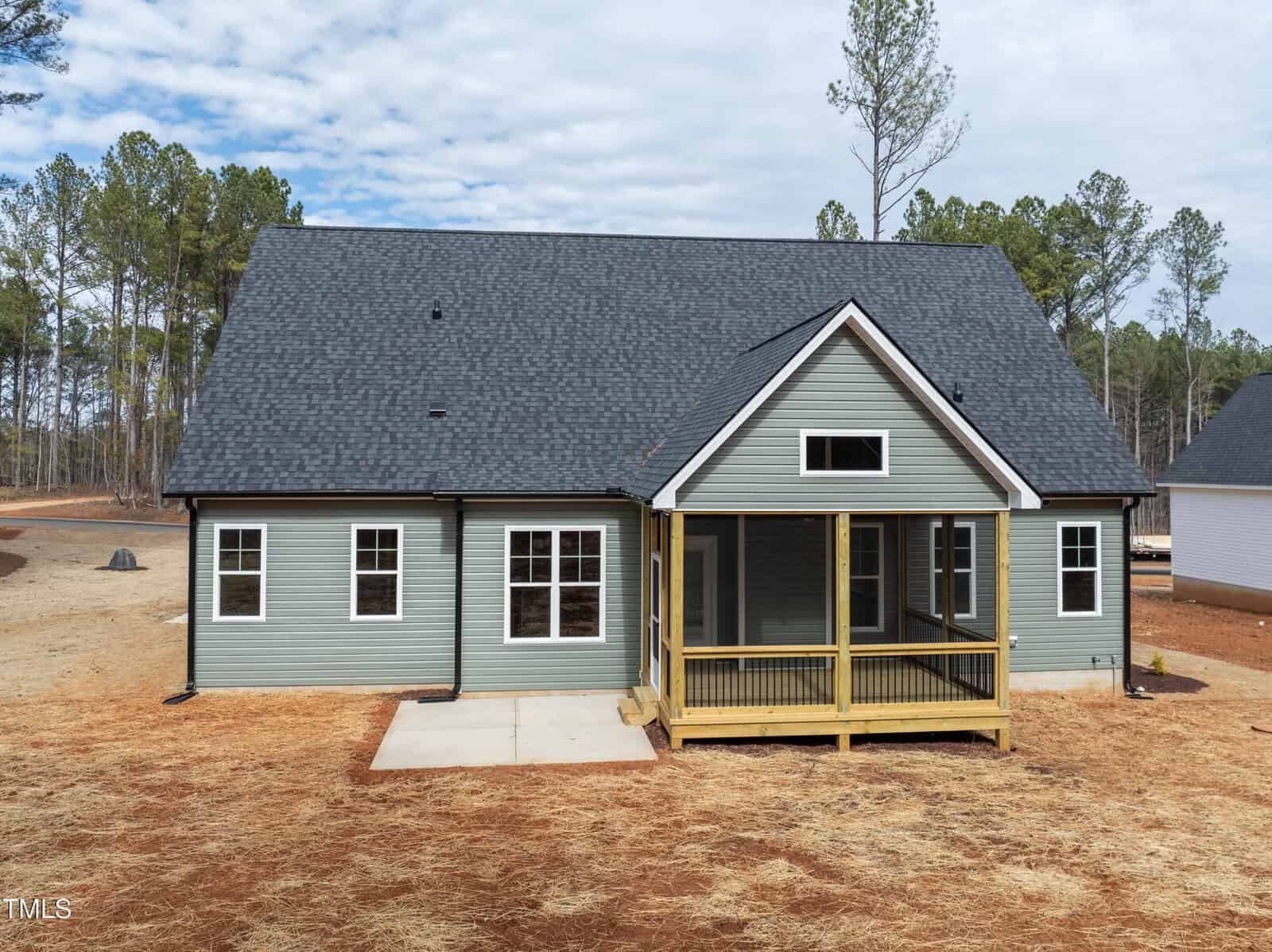 A newly built gray house with a dark roof, multiple windows, and a covered back porch, surrounded by bare ground and tall pine trees under a partly cloudy sky.