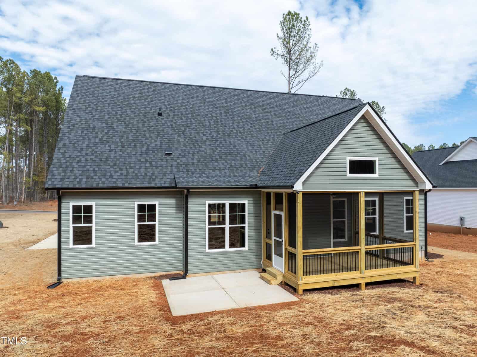 A new green house with black roof shingles, white-trimmed windows, a screened-in porch, and a concrete patio, surrounded by dry grass and tall trees in the background.