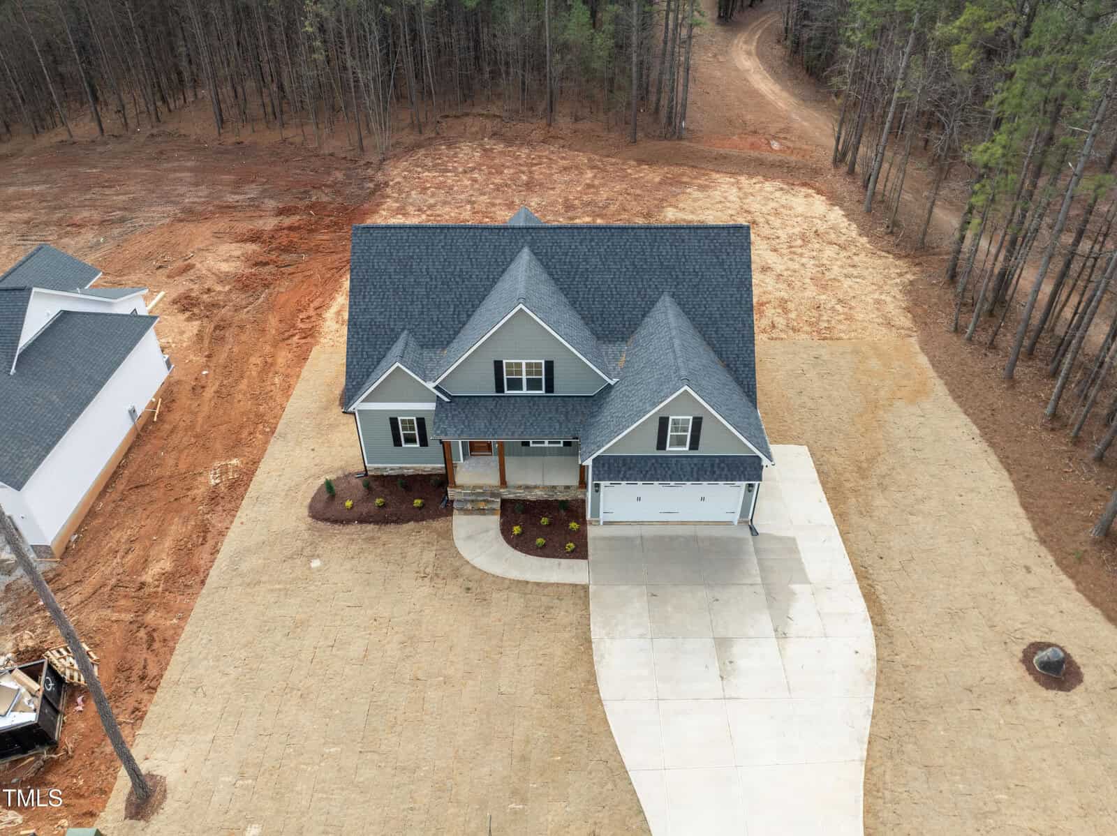 A two-story gray house with a covered front porch and attached two-car garage sits on a large, recently cleared plot with a concrete driveway, surrounded by trees and bare earth.