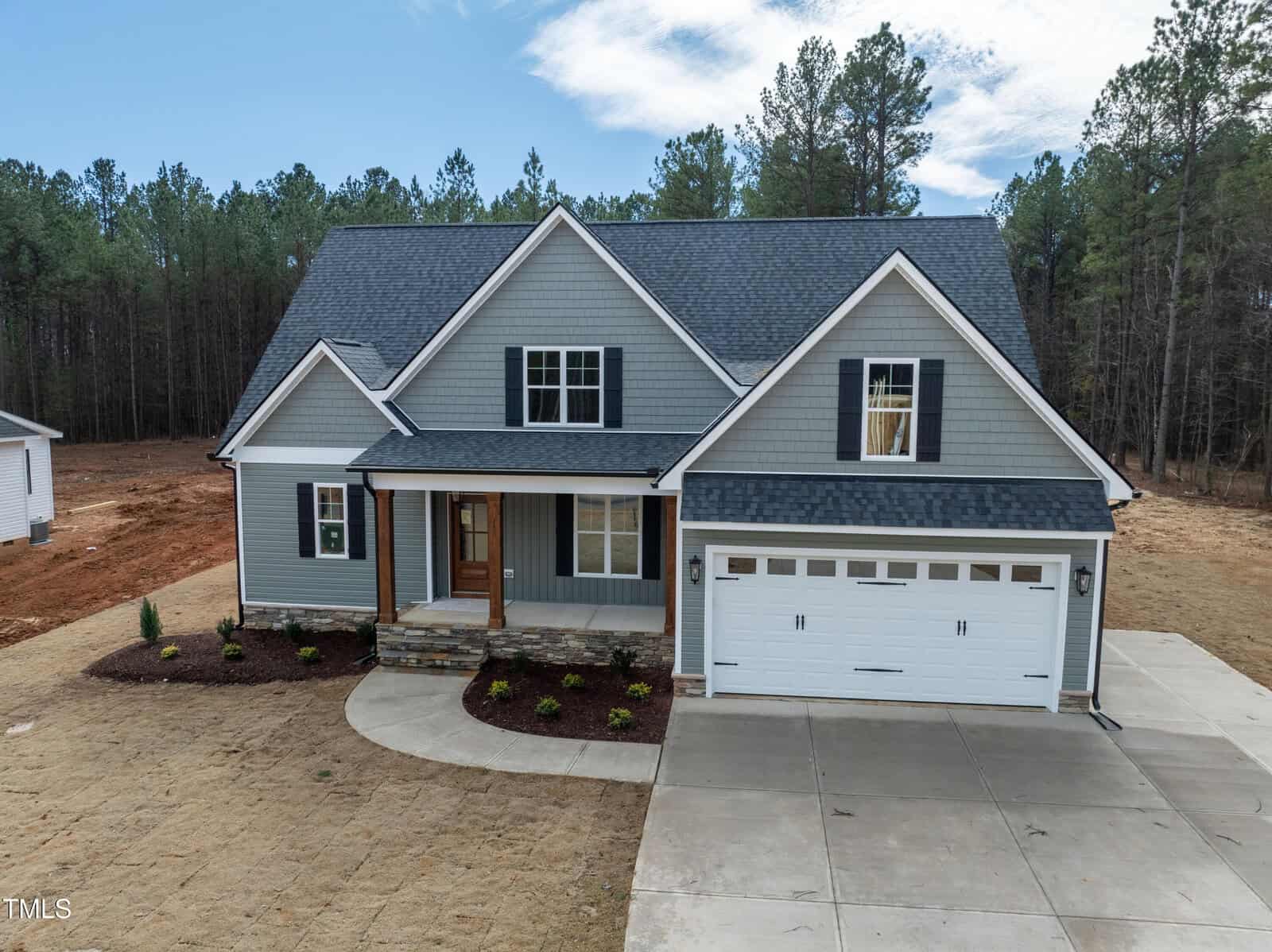 A two-story gray house with white trim, a covered front porch, and an attached two-car garage sits on a newly landscaped yard with mulched flower beds and a concrete driveway, surrounded by trees.