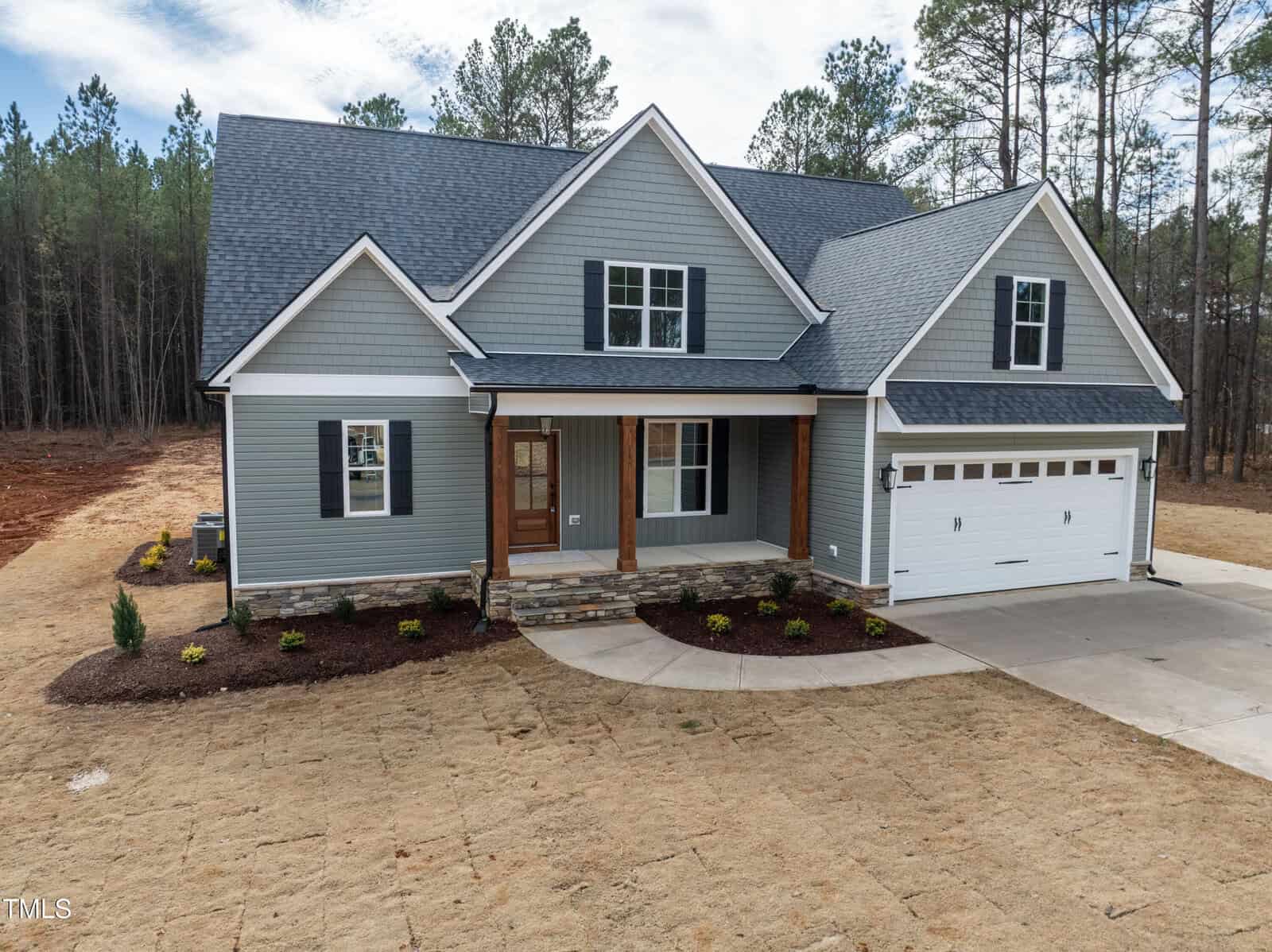 A gray two-story house with white trim and a covered front porch, featuring wooden columns, a stone base, and an attached two-car garage. The yard has minimal landscaping and new grass, with trees in the background.