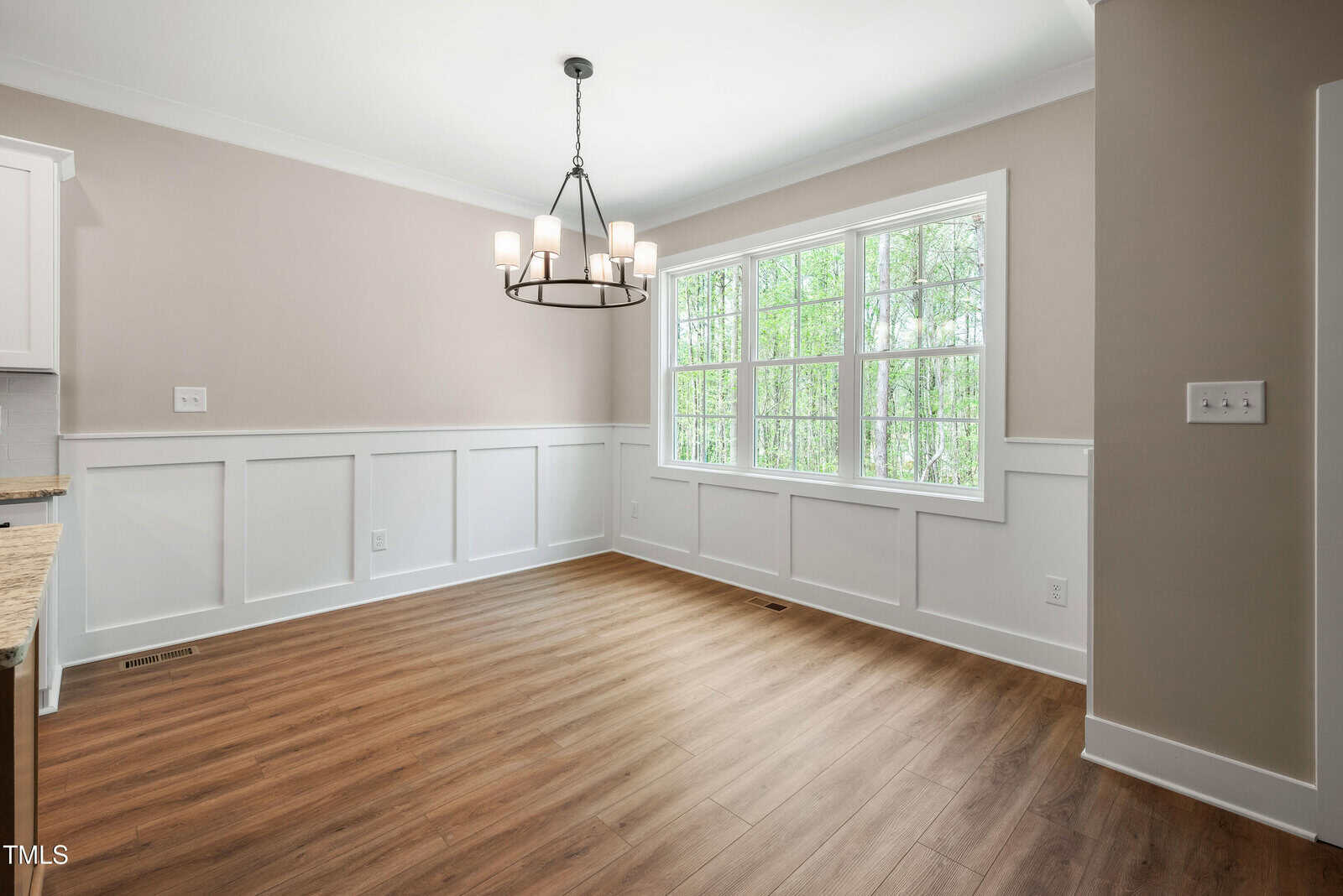 Bright dining area with large window, light wood flooring, beige and white paneled walls, and a black chandelier hanging from the ceiling. Natural light fills the room, and trees are visible outside the window.