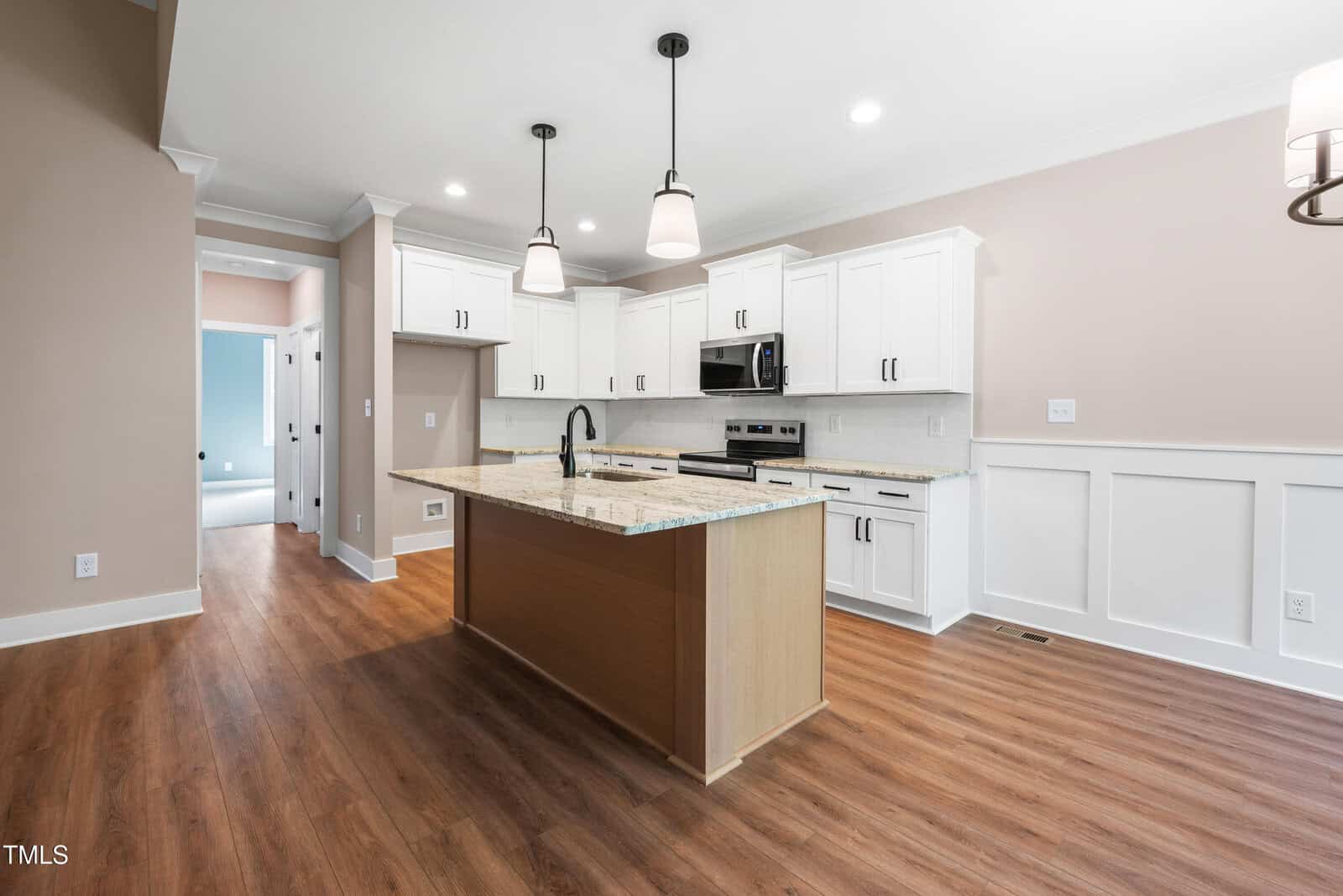 Modern kitchen with white cabinets, stainless steel appliances, a central island with sink, pendant lights, wood flooring, and beige walls with white trim. A hallway leads to other rooms.