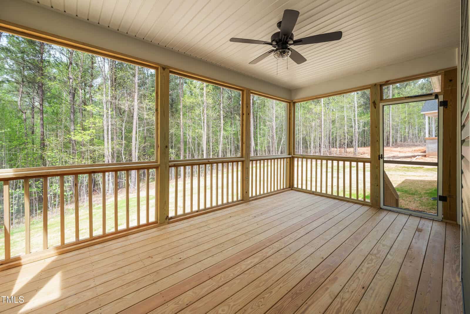A screened-in wooden porch with a ceiling fan, surrounded by forested trees. The porch has wooden railings, a door leading outside, and natural light coming through the screens.