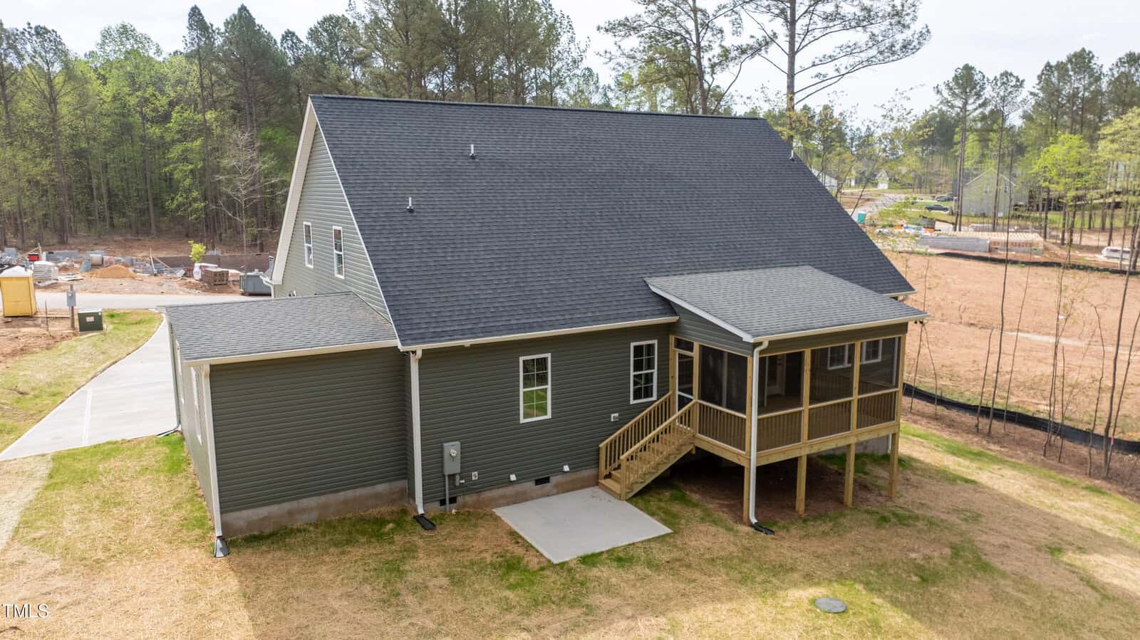 A gray, single-story house with a dark shingle roof, small covered porch, screened-in patio, and wooden steps. The backyard has patchy grass and is bordered by trees, with construction visible in the background.