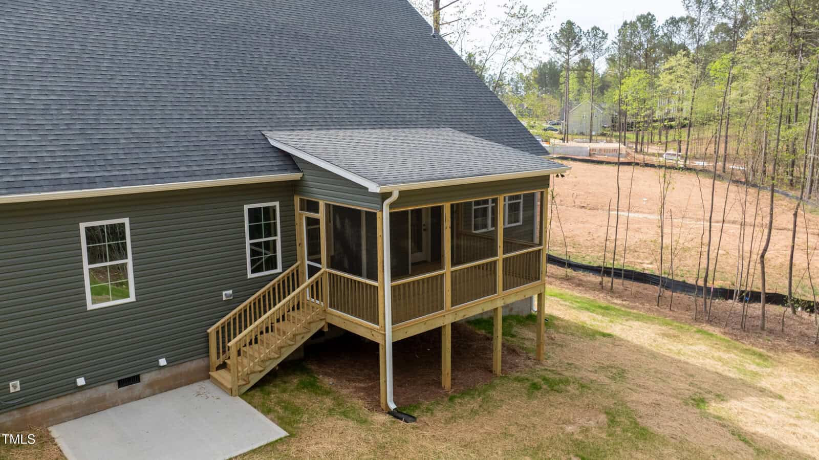 A green house with gray roof shingles features a screened-in porch elevated on wooden posts, accessed by a small staircase. The backyard has grass, trees, and a partly cleared area with a visible black silt fence.
