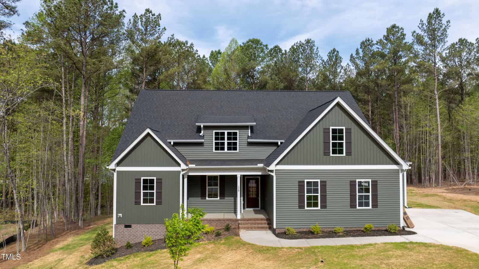 A two-story house with dark gray siding and white trim, a covered front porch, and a black shingled roof. The home is surrounded by tall trees, with a concrete driveway and minimal landscaping in front.