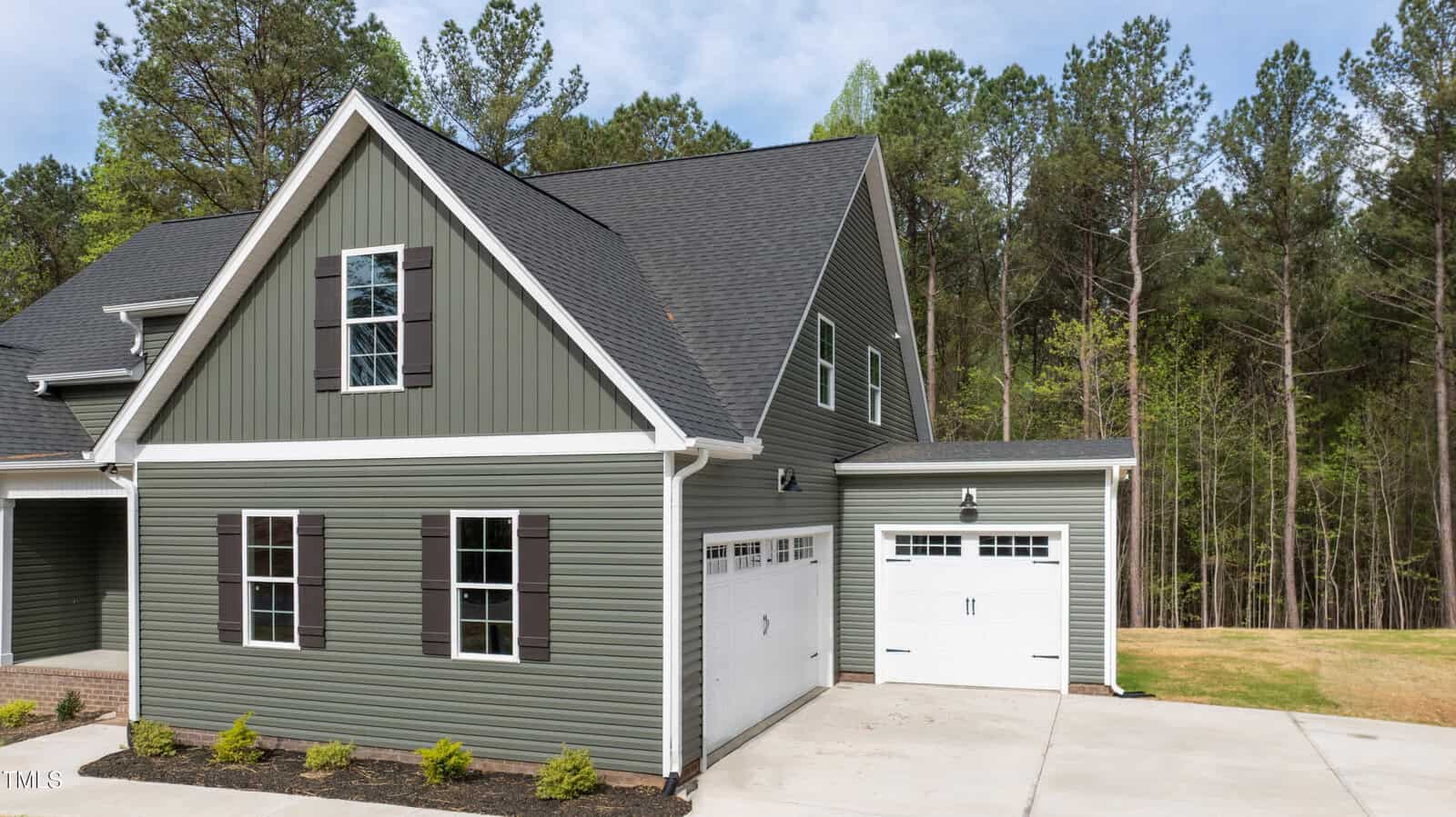 A green two-story house with black shutters, a black roof, two white garage doors, and a concrete driveway, set in front of a wooded area with tall trees under a partly cloudy sky.