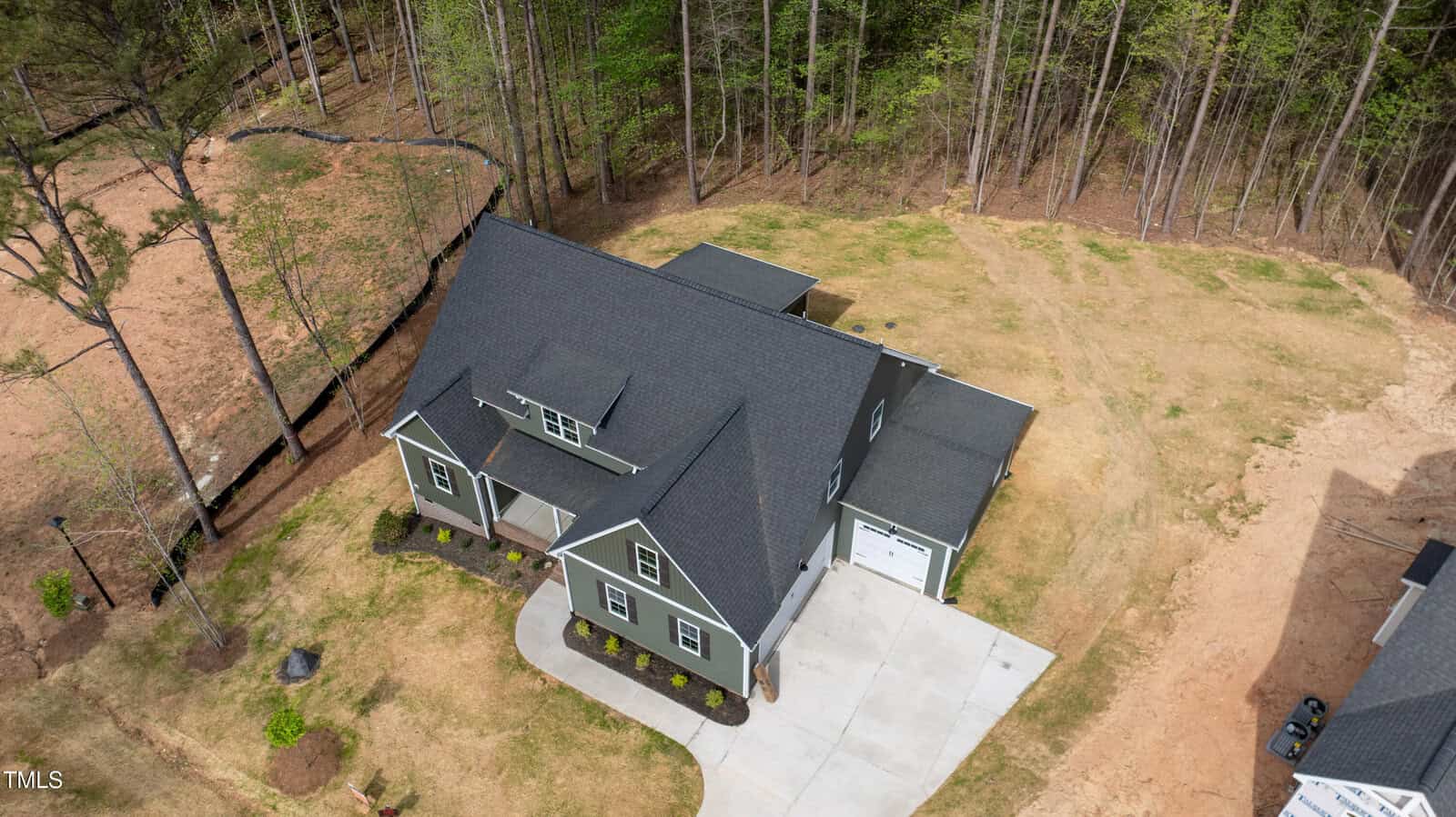 Aerial view of a gray two-story house with white trim, a covered front porch, and attached garage, surrounded by a mostly bare yard with some grass and trees at the edge of the property.