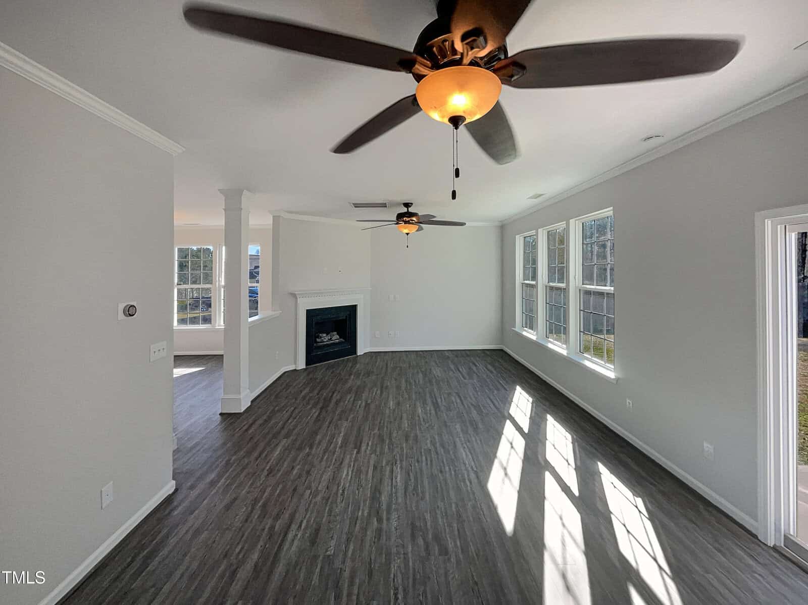 Spacious, empty living room with large windows letting in natural light, wood flooring, white walls, and a ceiling fan with lights. A fireplace is in the far corner, and an adjoining hallway is visible on the left.