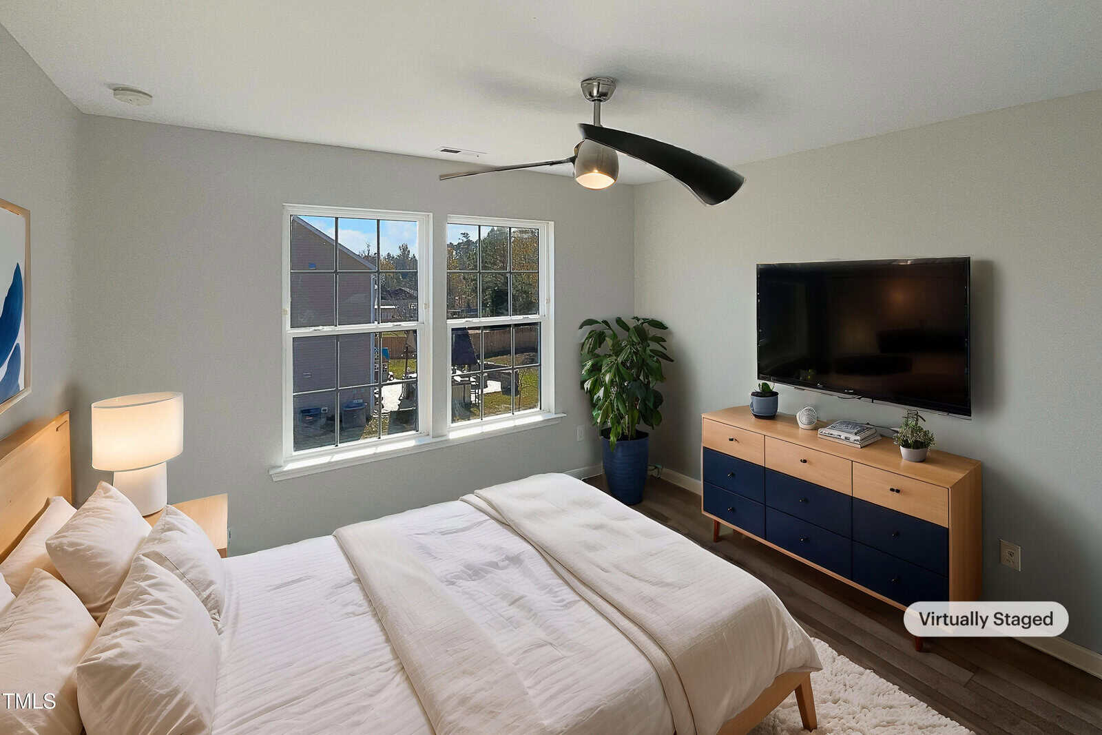 A bedroom with a bed, dresser, and TV. The bed has white bedding, and a large window lets in natural light. A plant is in the corner, and a ceiling fan hangs above. The room is virtually staged.