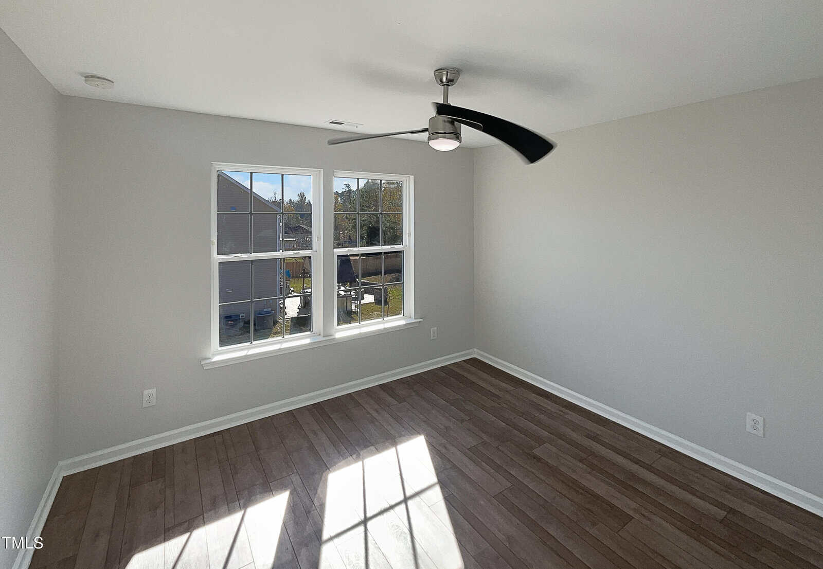A bright, empty room with light gray walls, a ceiling fan, and two large windows. The floor is wood, and sunlight casts a rectangular pattern on it. The windows offer a view of a neighboring house and some trees.