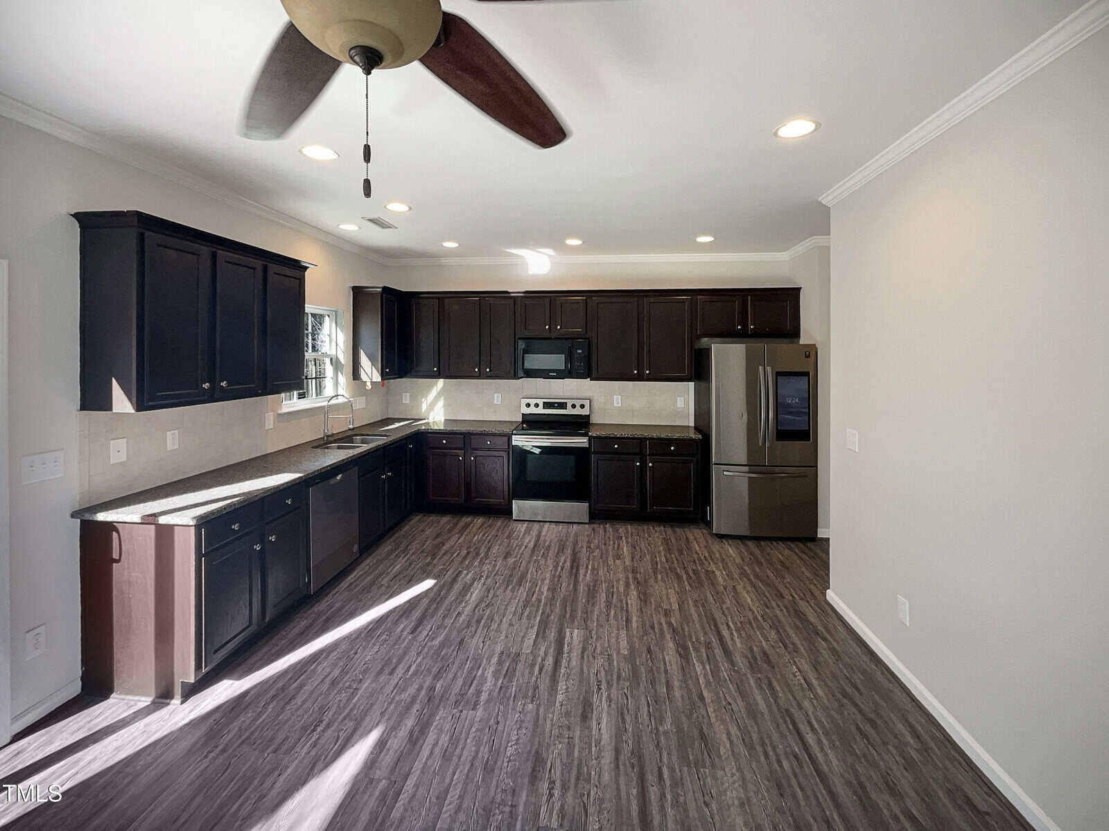 A kitchen with dark wood cabinets, stainless steel appliances, and a light wood floor. The room features a ceiling fan, recessed lighting, and sunlight streaming in from a window on the left. The walls are painted light gray.
