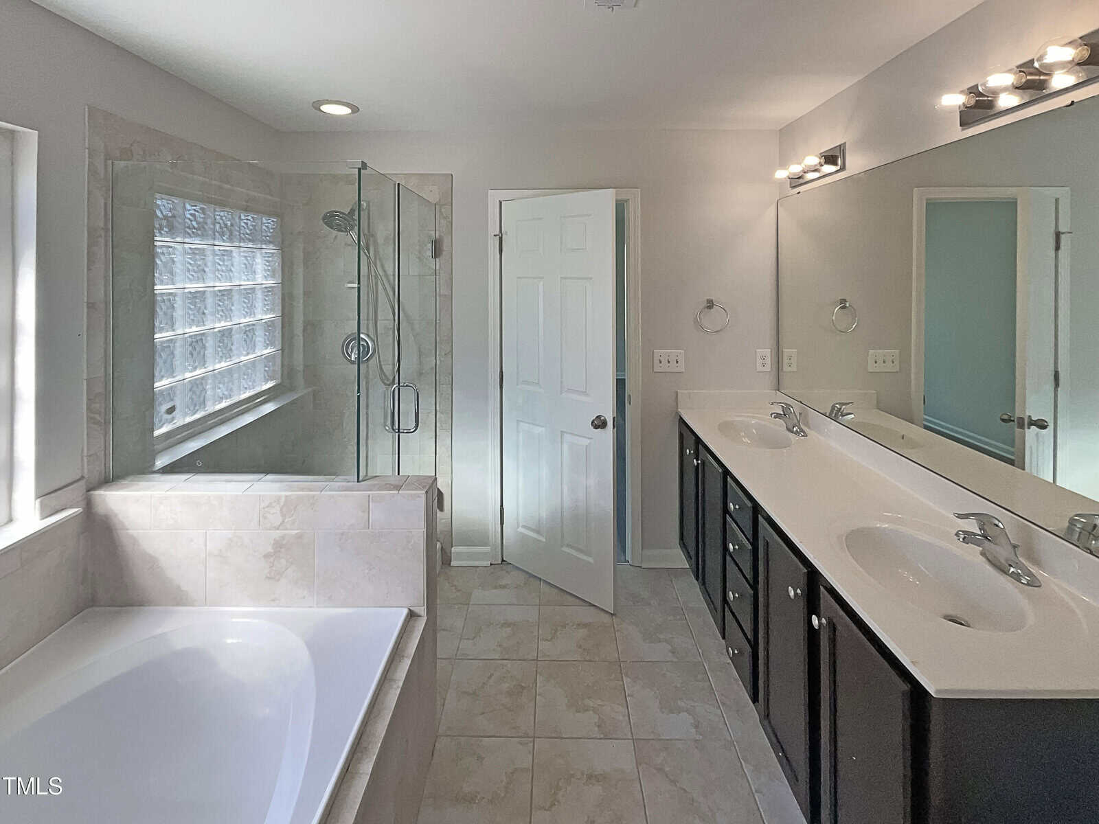A modern bathroom featuring a large bathtub, a glass-enclosed shower, and a double sink vanity. The room has tiled floors and a window with frosted glass blocks. The walls are light-colored with a door leading to an adjacent space.
