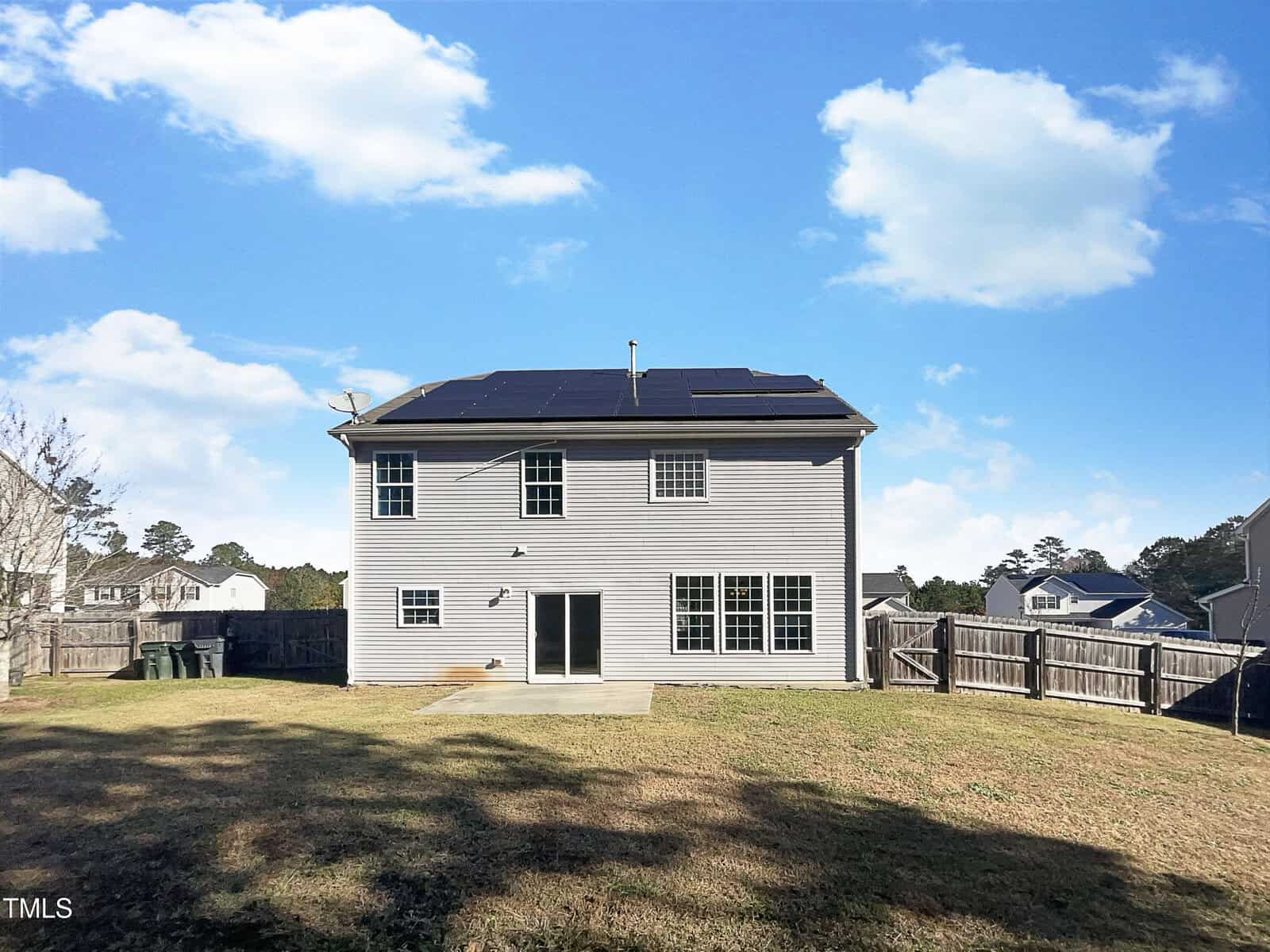A two-story house with solar panels on the roof is pictured on a clear day. The house has a beige exterior and multiple large windows. A fenced backyard with grass surrounds the property. Several neighboring houses are visible in the background.