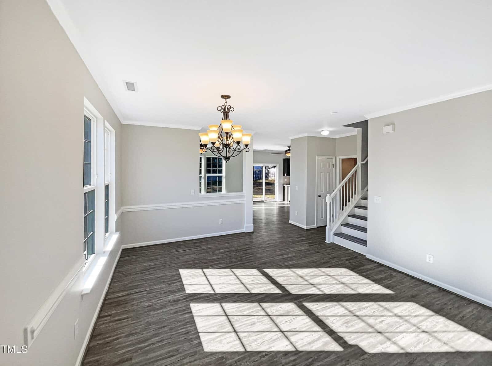 A spacious, unfurnished living room with large windows letting in natural light. It features light gray walls, dark wood flooring, a decorative chandelier, and white trim. A staircase and a doorway lead to other parts of the house.