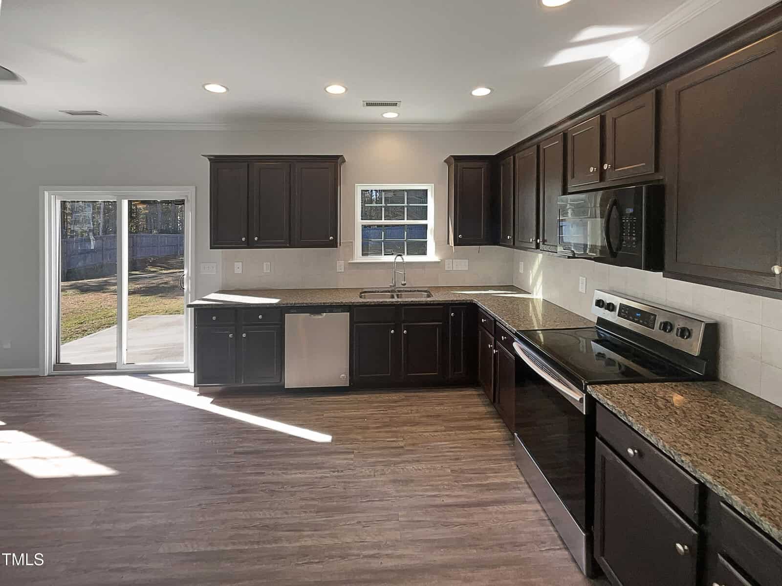 A modern kitchen with dark wood cabinets and stainless steel appliances, including a stove, microwave, dishwasher, and sink. The room features a tiled backsplash, light countertops, and large sliding glass doors leading to an outdoor area.
