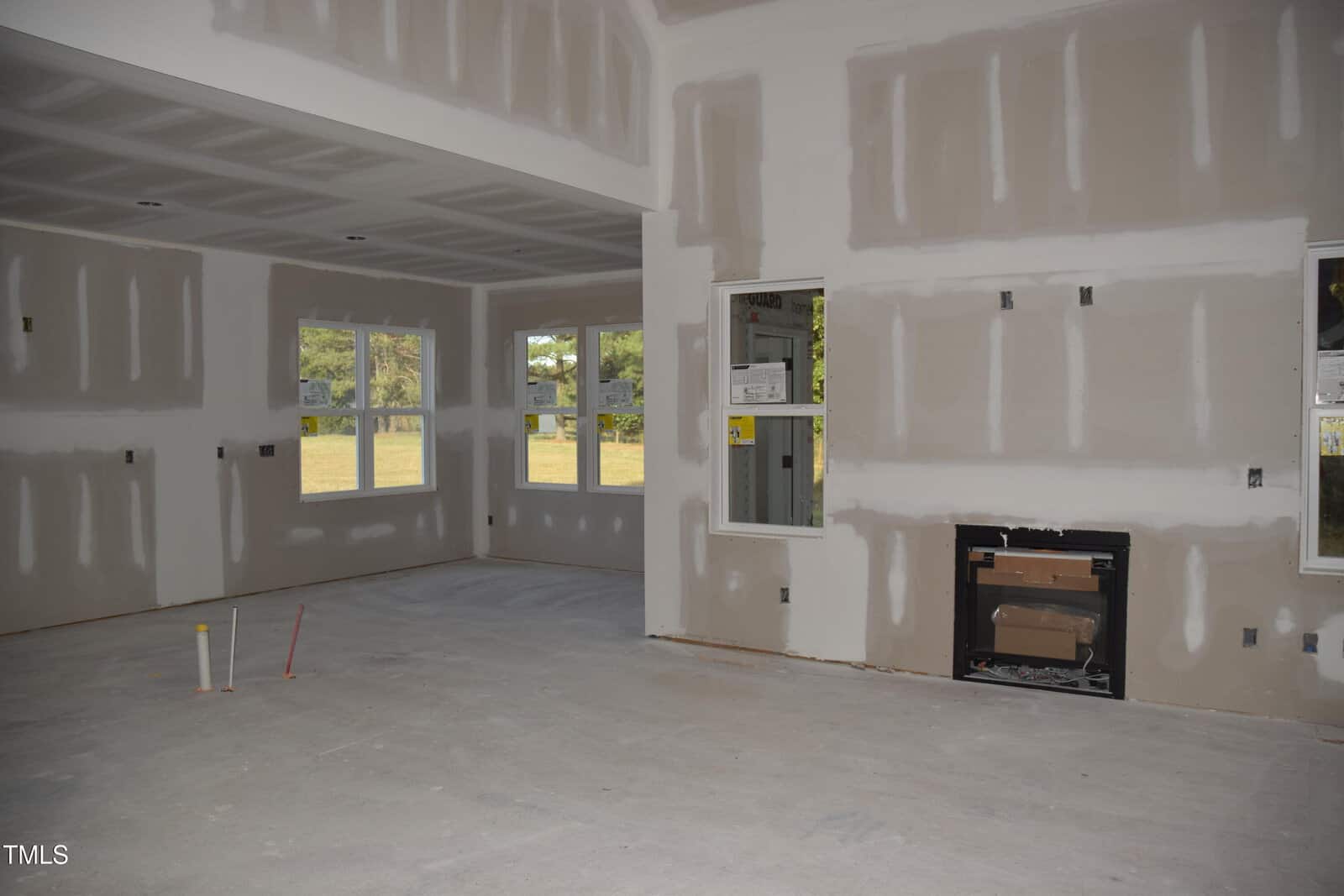 Interior view of a partially constructed room with unfinished drywall, exposed electrical outlets, and plumbing fixtures. There are large windows with a view of trees outside. A fireplace area is framed but uncompleted, and the floor is bare concrete.