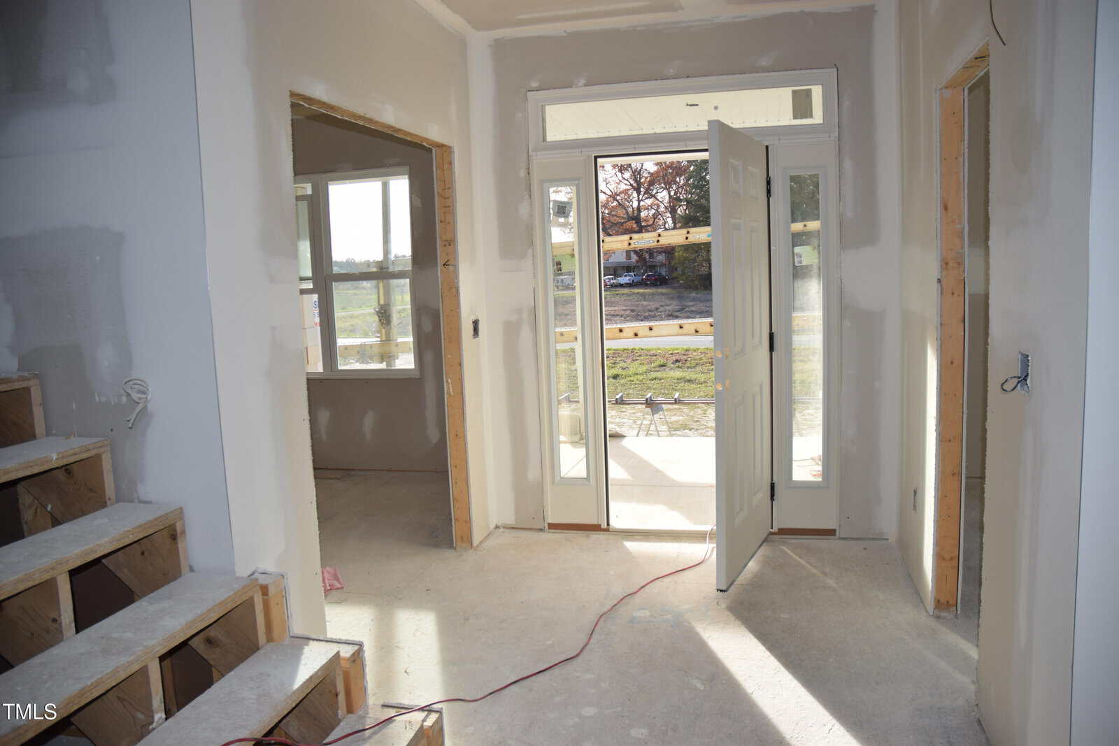 A partially completed interior of a house, featuring a white front door slightly open, exposing a view outside. Unfinished drywall and wooden staircase are visible, along with construction materials on the floor. Electrical wiring is seen on the walls.