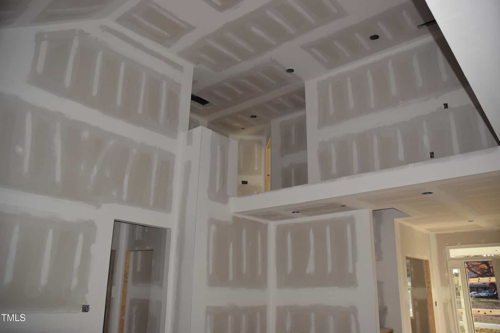 The image shows an interior view of a house under construction. The walls and ceiling are made of drywall panels, with visible joint tape and compound. Natural light enters through a window and a glass door on the right side.