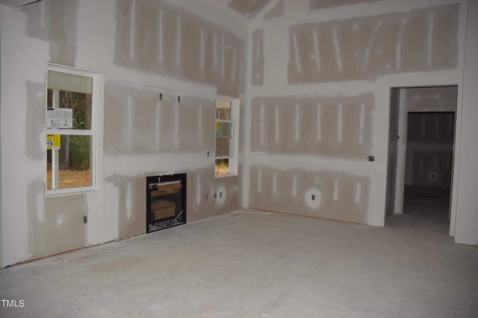 A room under construction with unpainted drywall on the walls and ceiling. There are two windows on one wall and a door frame on another. Electrical outlets are visible, and there is an uncovered floor.