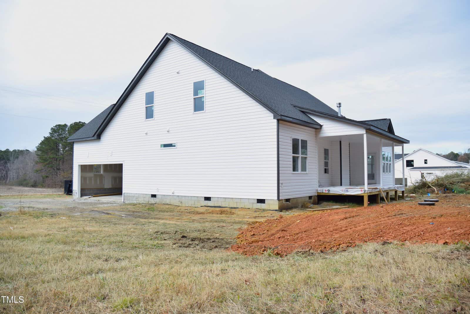 A newly constructed white house with a black roof sits on a grassy plot. It features a covered porch and a two-car garage. The surrounding area includes a patch of bare soil and a rural landscape with trees in the background.